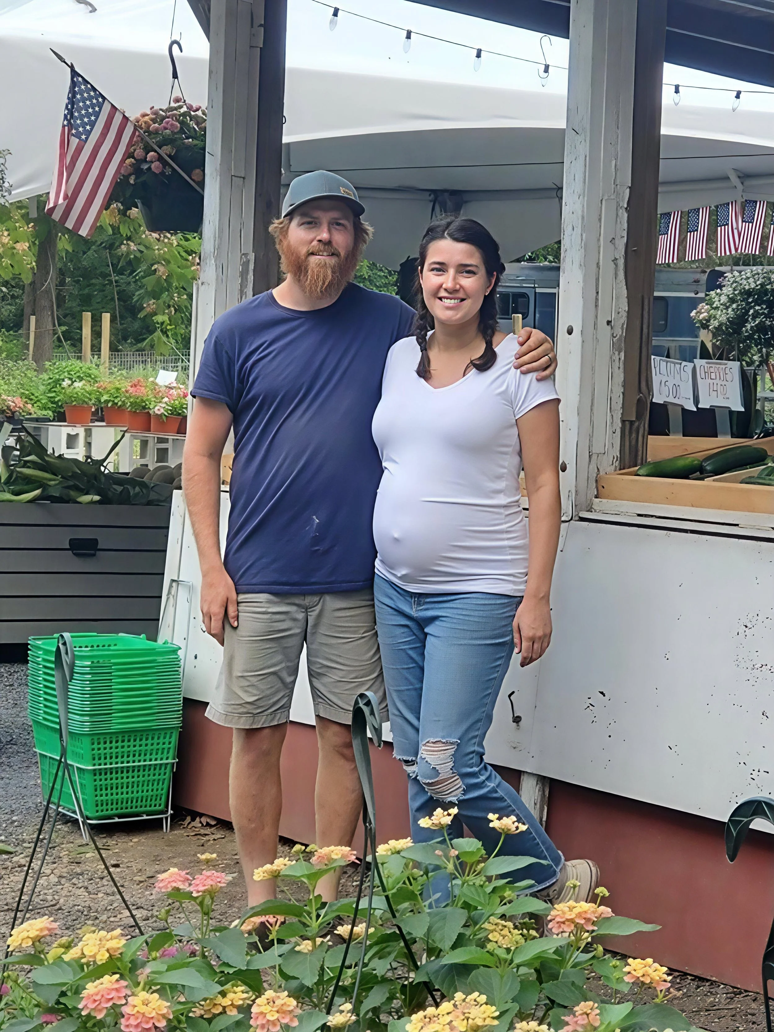 A smiling young woman with long dark hair in braids, wearing a white t-shirt and ripped jeans, standing next to a bearded man in a blue t-shirt and gray cap, at an outdoor farm or market stand with flowers and vegetables