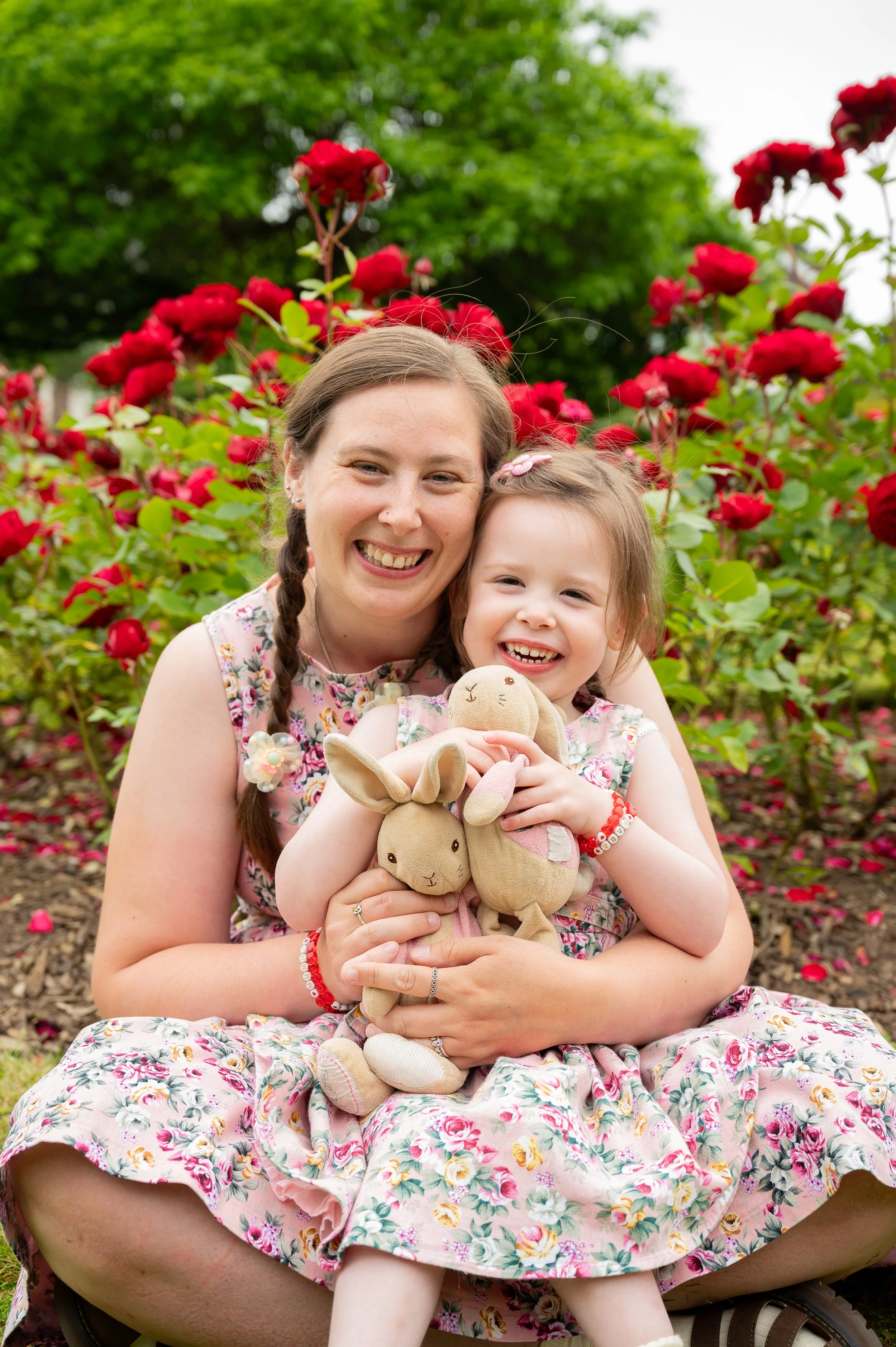 A girl and her mum smile for the camera. The image is colourful and both wear matching flowery dresses and the girl holds two cuddly bunny toys. Red flowers are visible in the background.