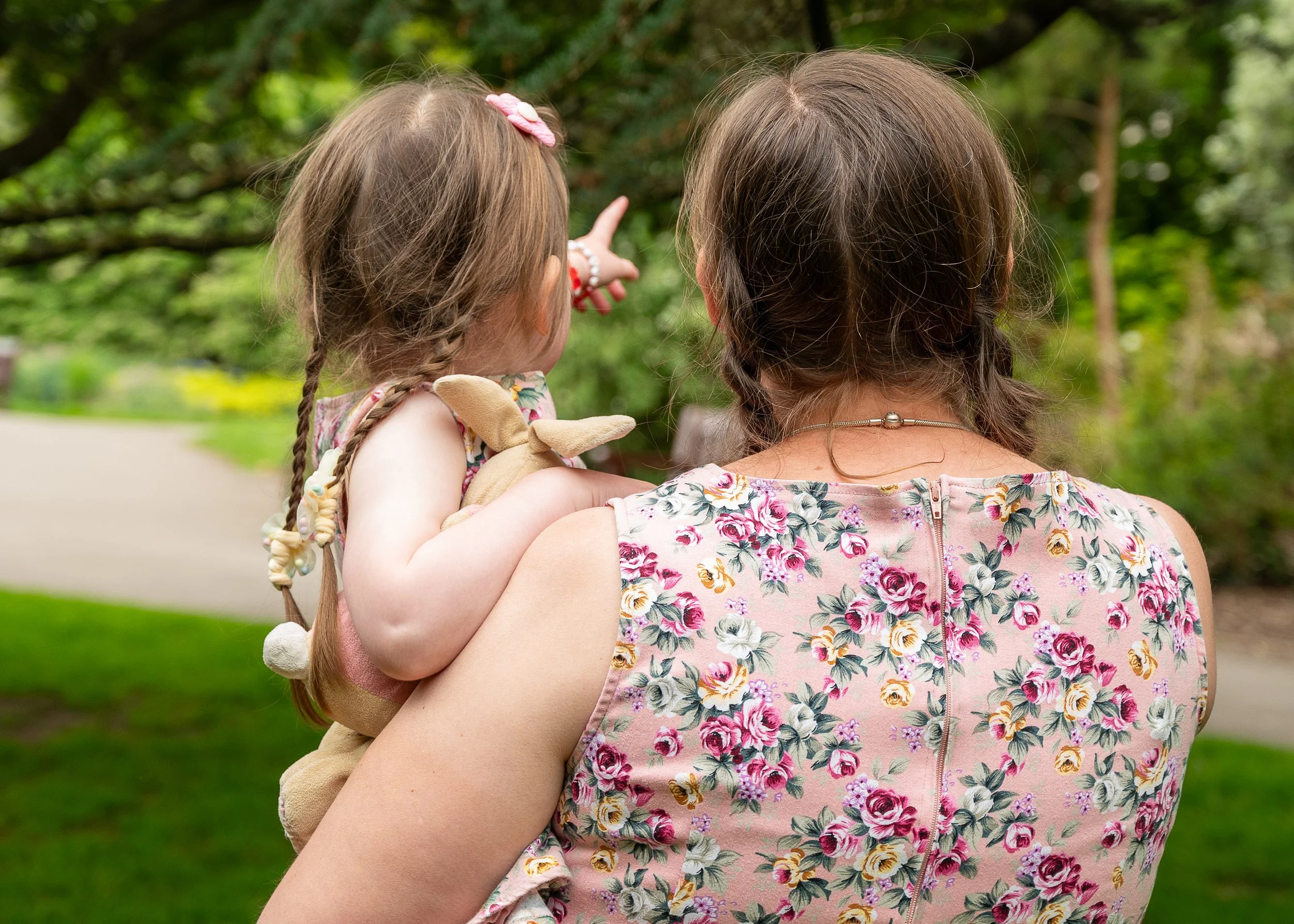 A girl holding a toy bunny faces away from camera being held by her mum. The image is colourful and both wear matching flowery dresses.