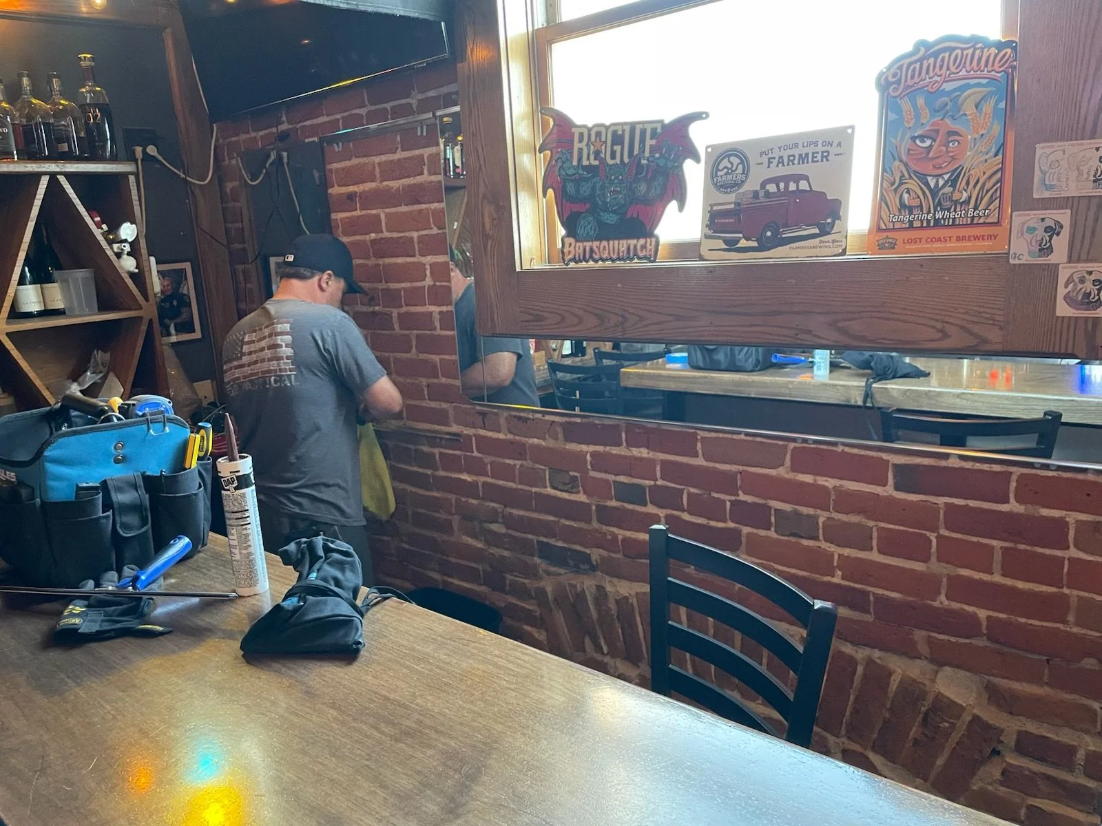A person wearing a black cap and gray t-shirt standing near a brick wall, with tools and a bag on the table in the foreground, and a mirror reflecting part of his back. The wall has lighthouse and beer posters, and there is a window with more signs a