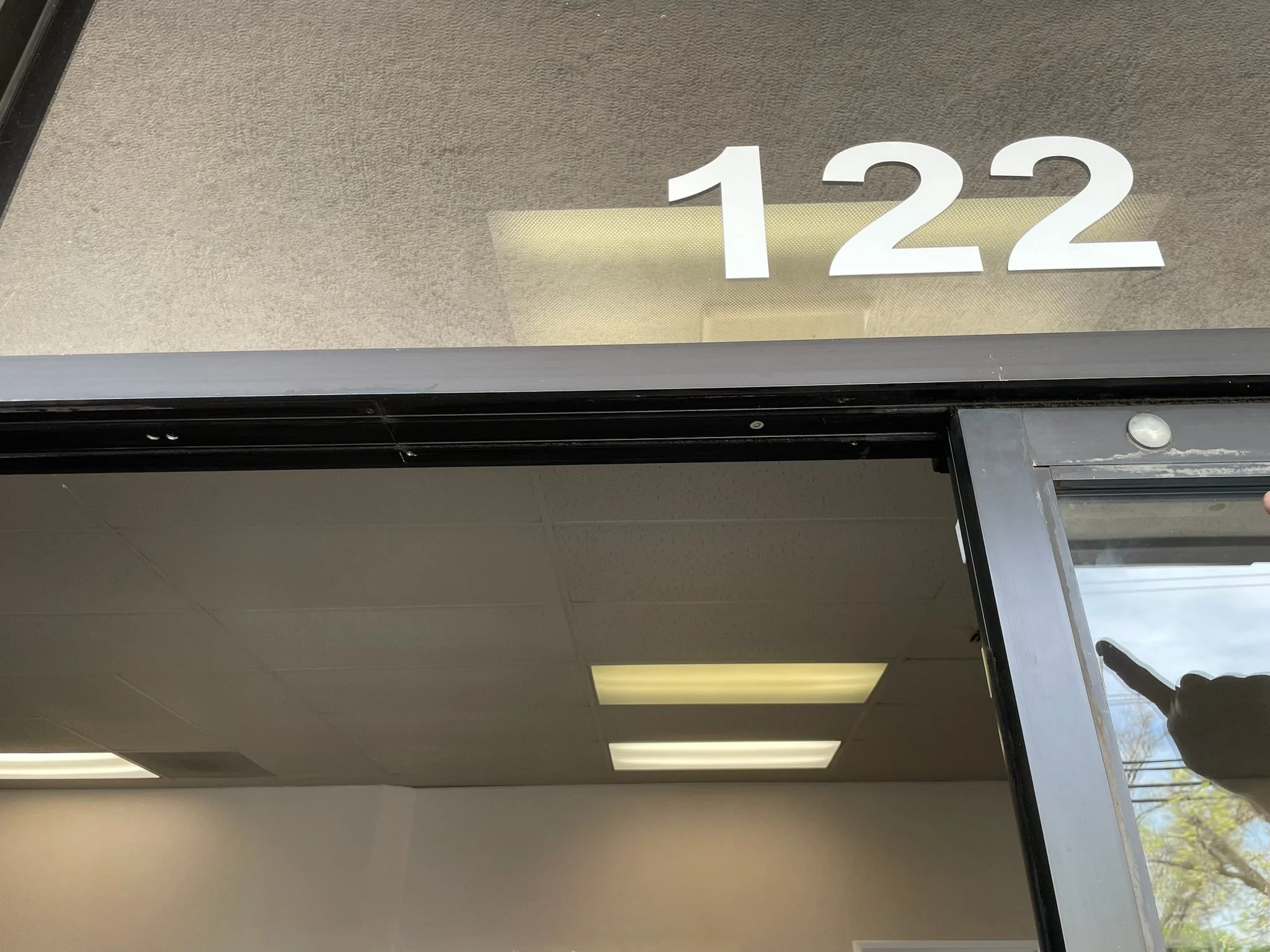 Door with the number 122 on the glass window above it, showing an indoor ceiling with fluorescent lights and part of a person's hand holding a phone or camera in the reflection.