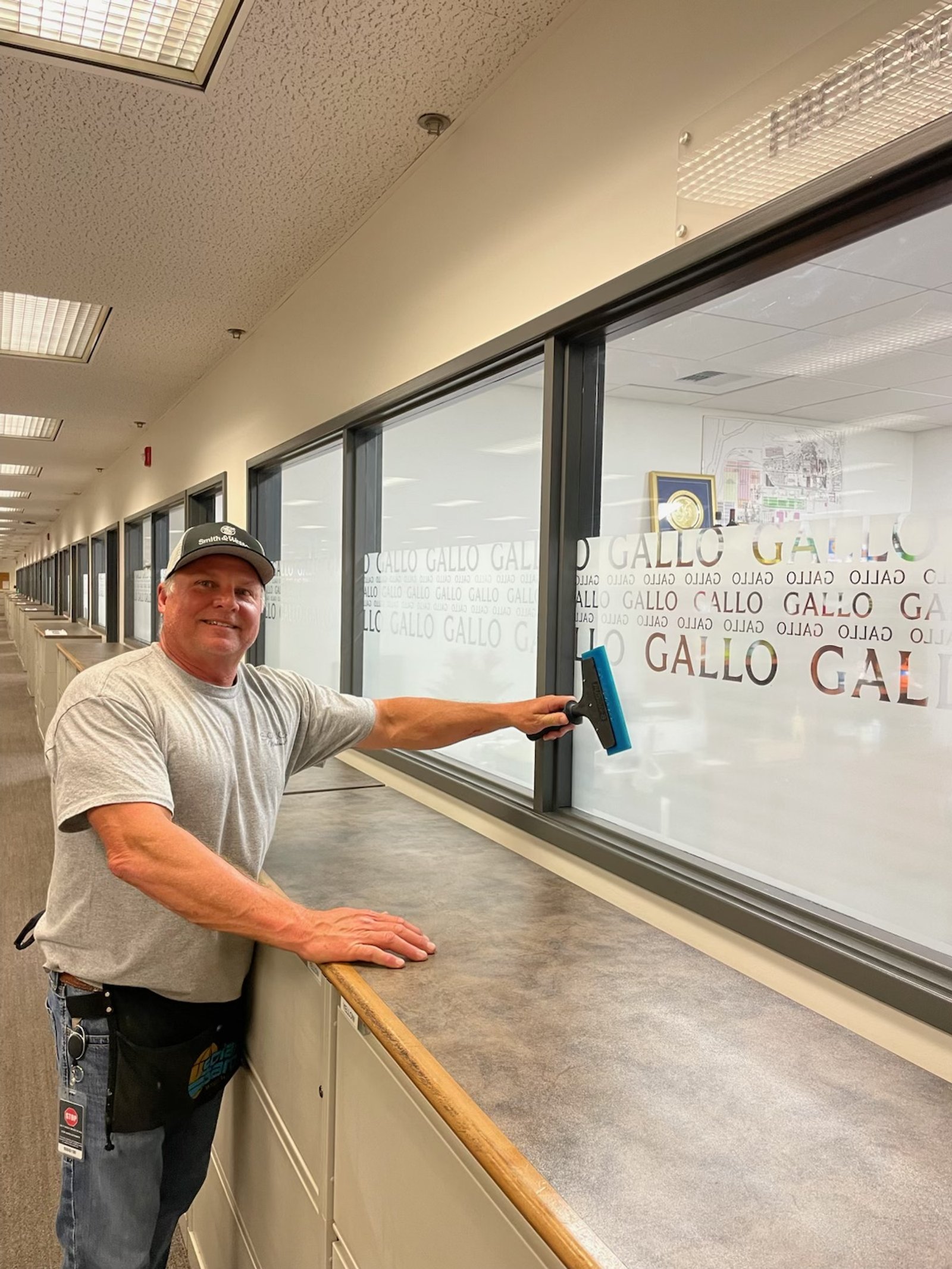 A man cleaning a large glass window in an indoor hallway while smiling at the camera.