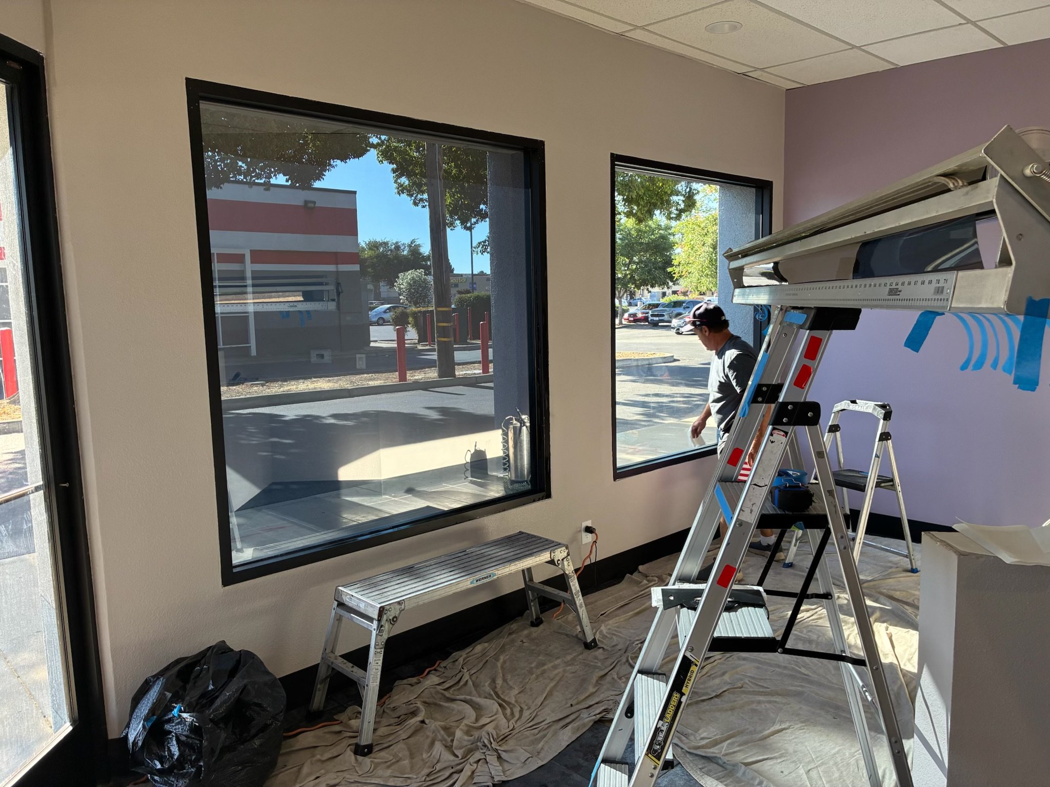 A worker installing large window panes in a room with light-colored walls and a purple accent wall. The room has two large windows, aluminum ladders, a step stool, and protective coverings on the floor.