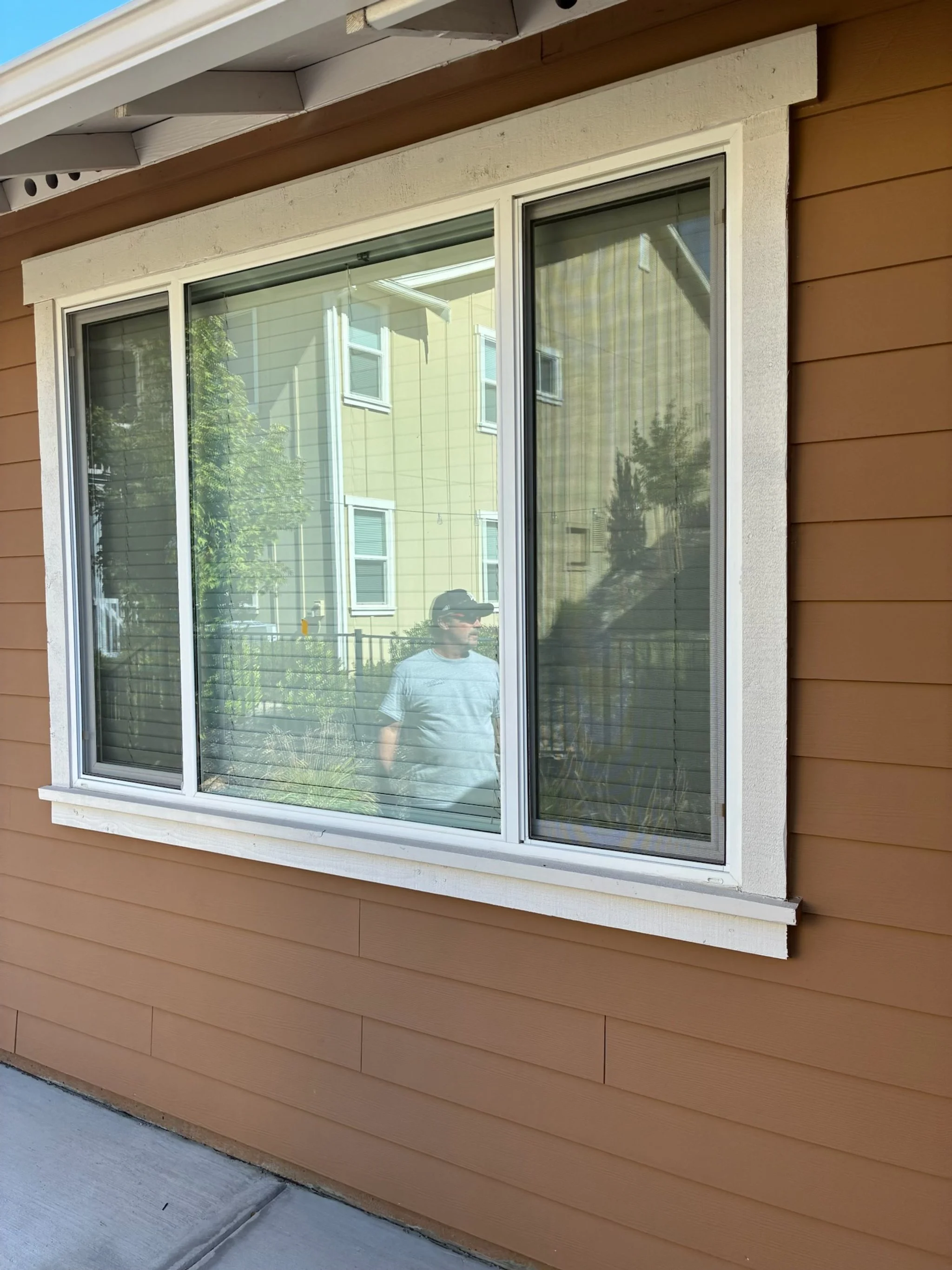 Exterior of a house with a large window reflecting a person standing outside, with neighboring houses and greenery visible in the reflection.