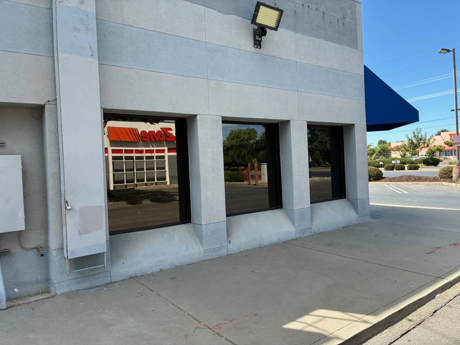 Side of a commercial building with large windows, a security light, and a blue awning, with a parking lot and trees in the background.