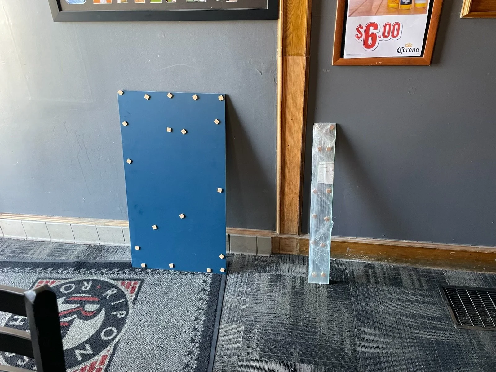 A blue vertical climbing wall panel with small white holds leaning against a gray wall. To the right, there is a tall, metal climbing hold with multiple bolt holes. Part of a black and red floor mat with the logo 'Papa Ro' and a black chair leg are v