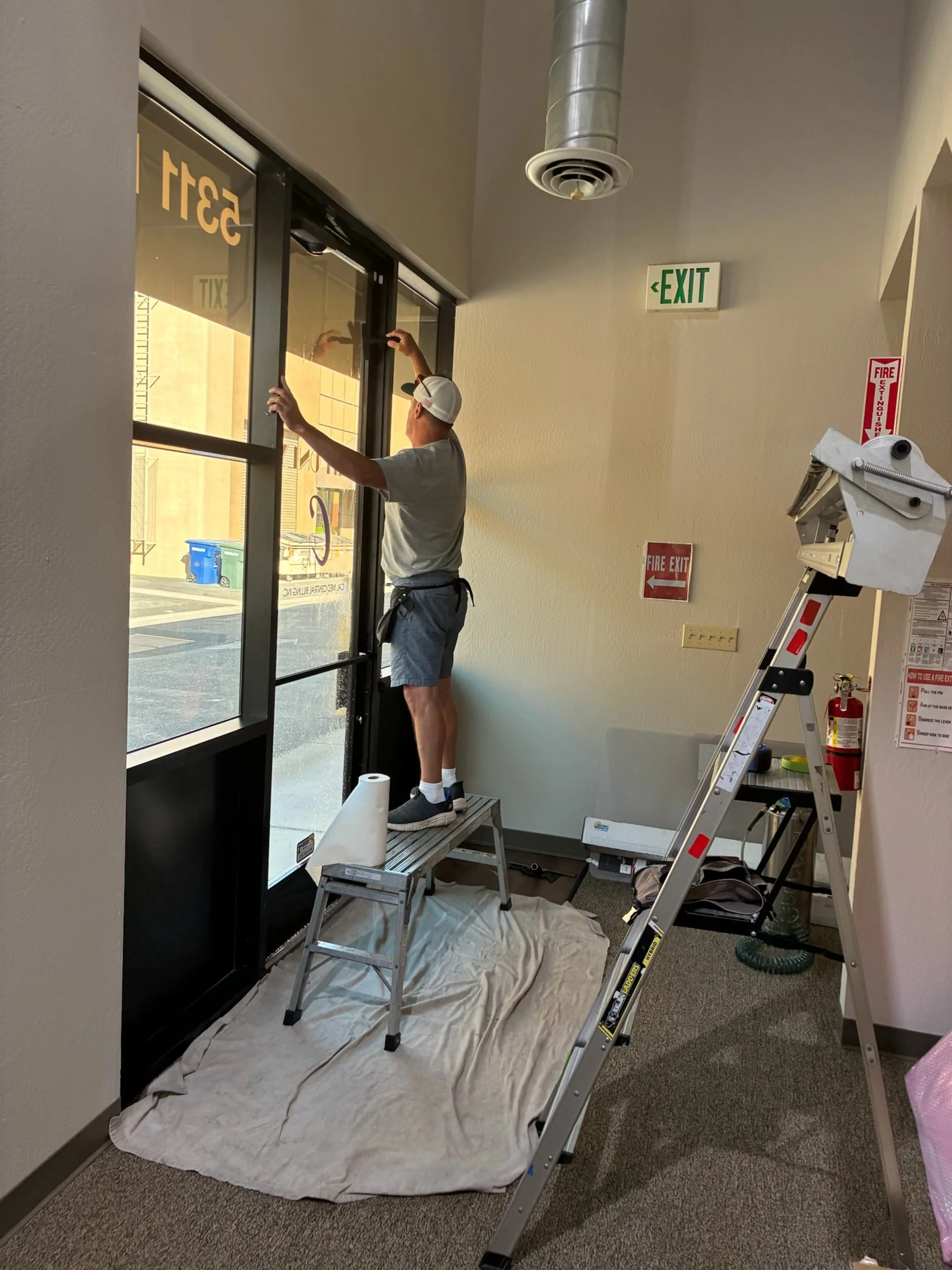 A man standing on a step ladder cleaning large glass windows at the entrance of a building.