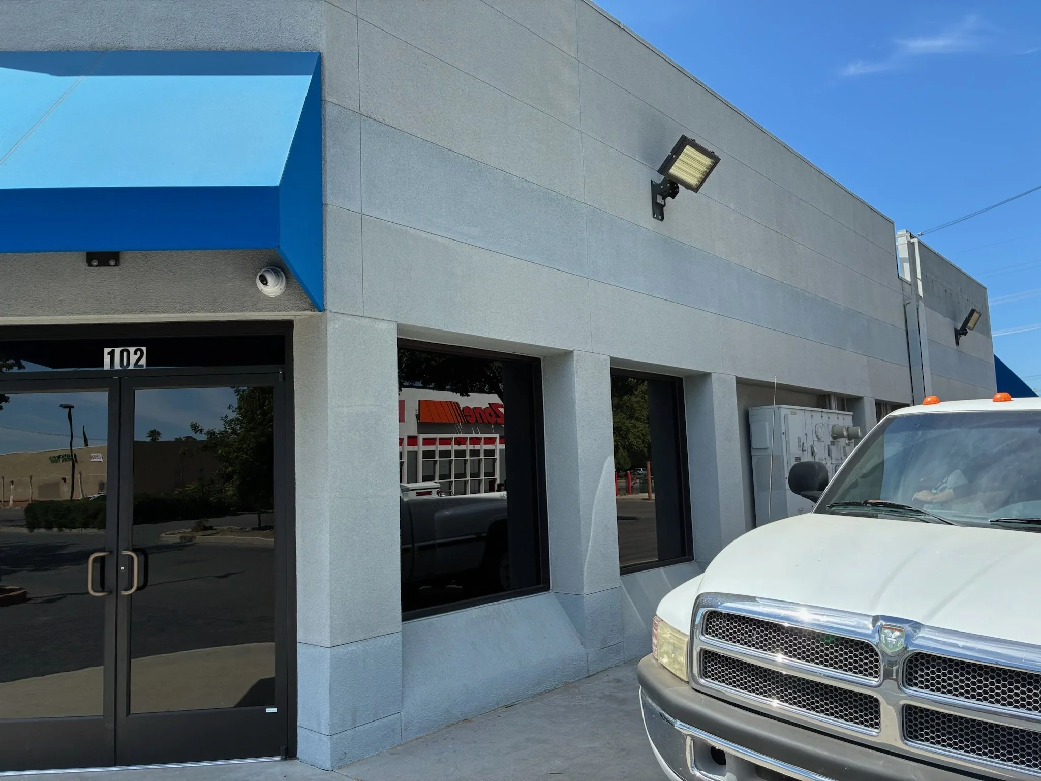 Exterior of a commercial building with large windows, a dark glass door, and a blue awning; a white van is parked in front. The building has security cameras and exterior lights, with a blue sky overhead.
