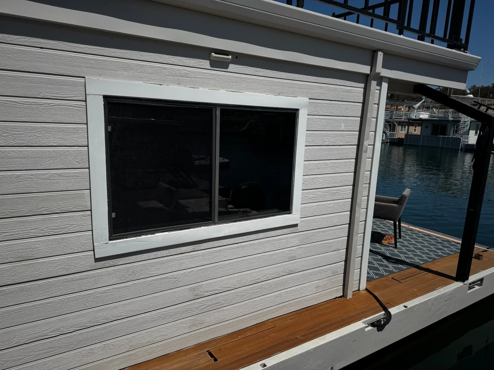 Close-up of a white houseboat with a window, on a dock by the water, with other boats and houseboats visible in the background.