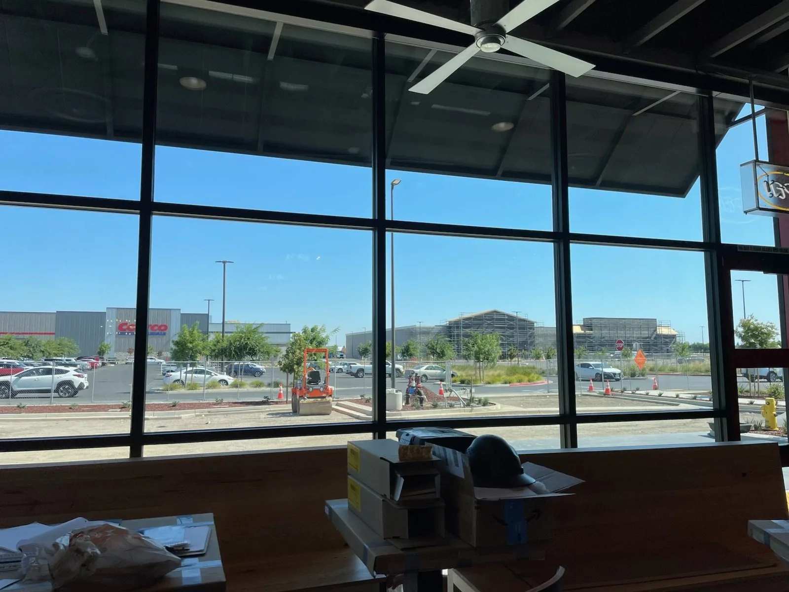 View through large glass window of a parking lot outside a shopping center, with cars, trees, and construction in the distance, under a clear blue sky.