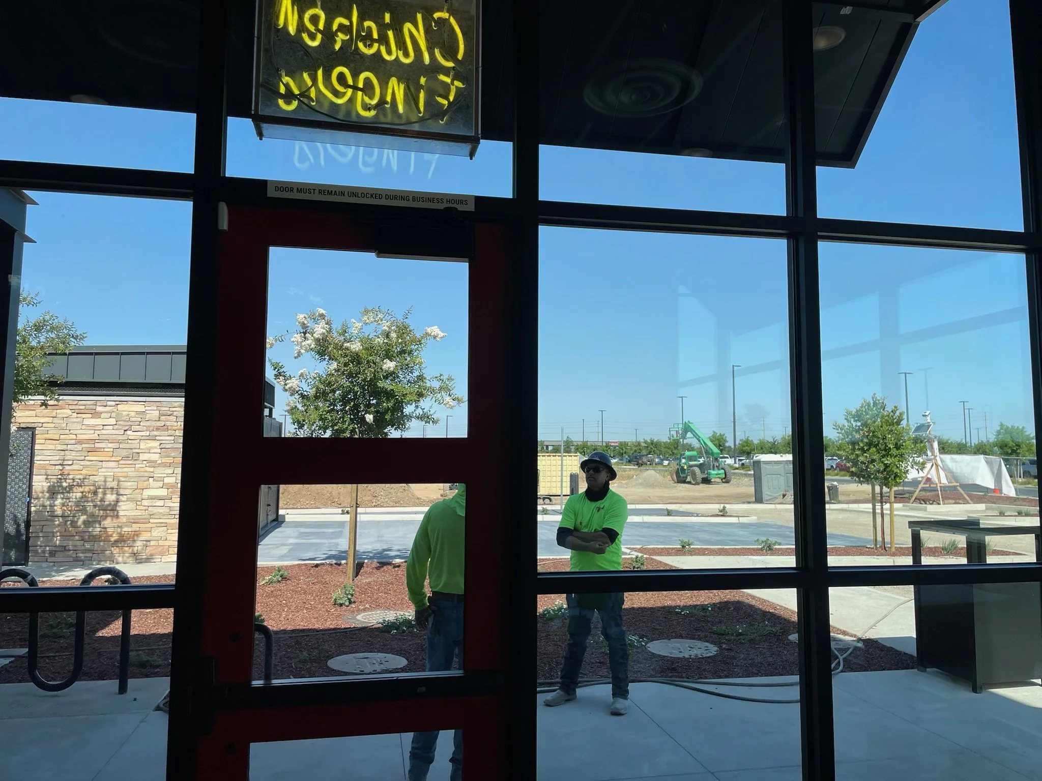 Inside view of a restaurant or cafe with a glass door and window, showing two workers outside wearing bright green shirts, one with arms crossed, during a sunny day.