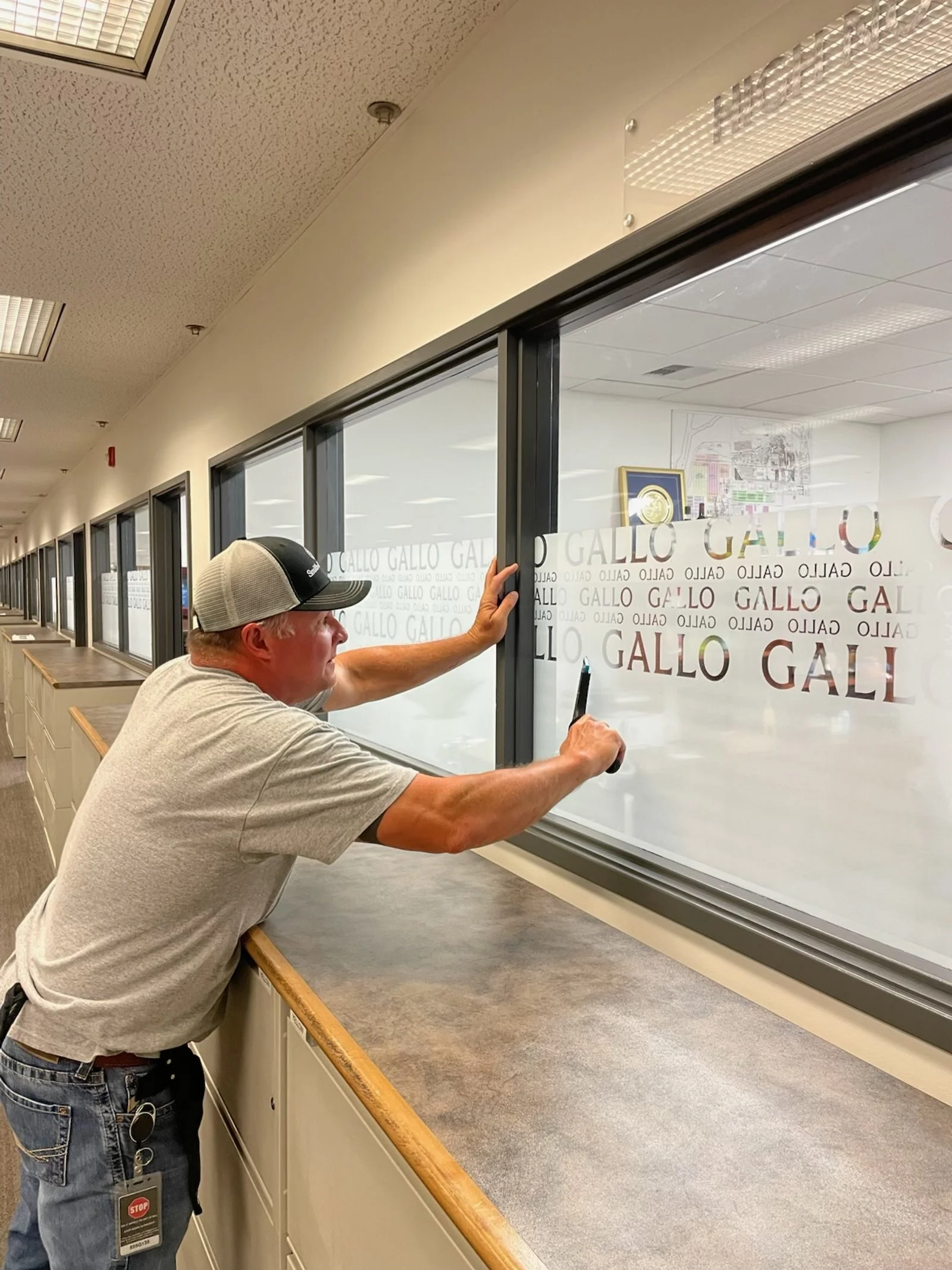 A man is applying vinyl lettering to a glass window in an office or building hallway. The window displays the name 'Gallo' multiple times.