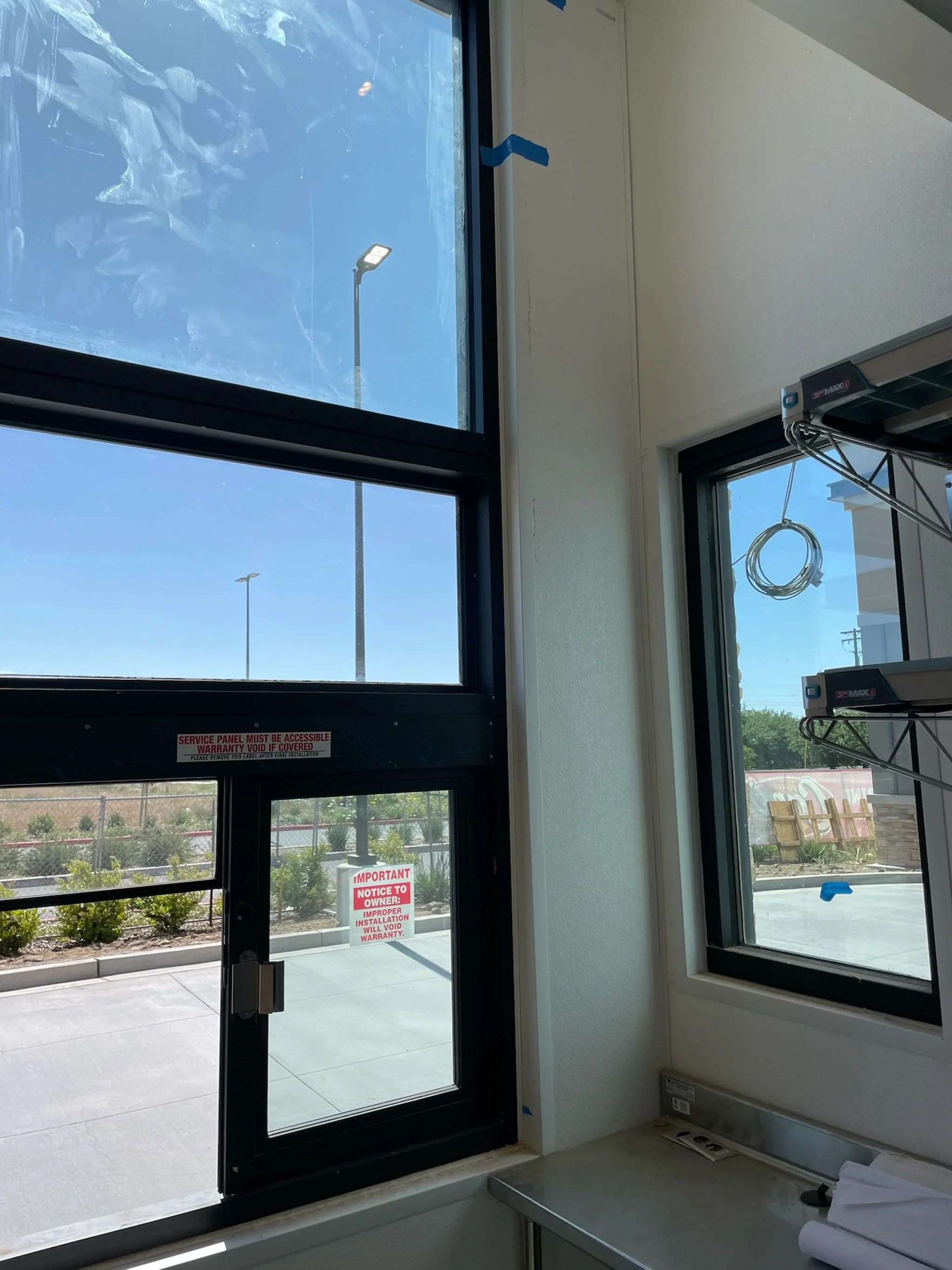 Interior view of a commercial building with large windows, showing a parking lot outside, some signs on doors, a shelf with coiled cables, and a metal countertop near the window.
