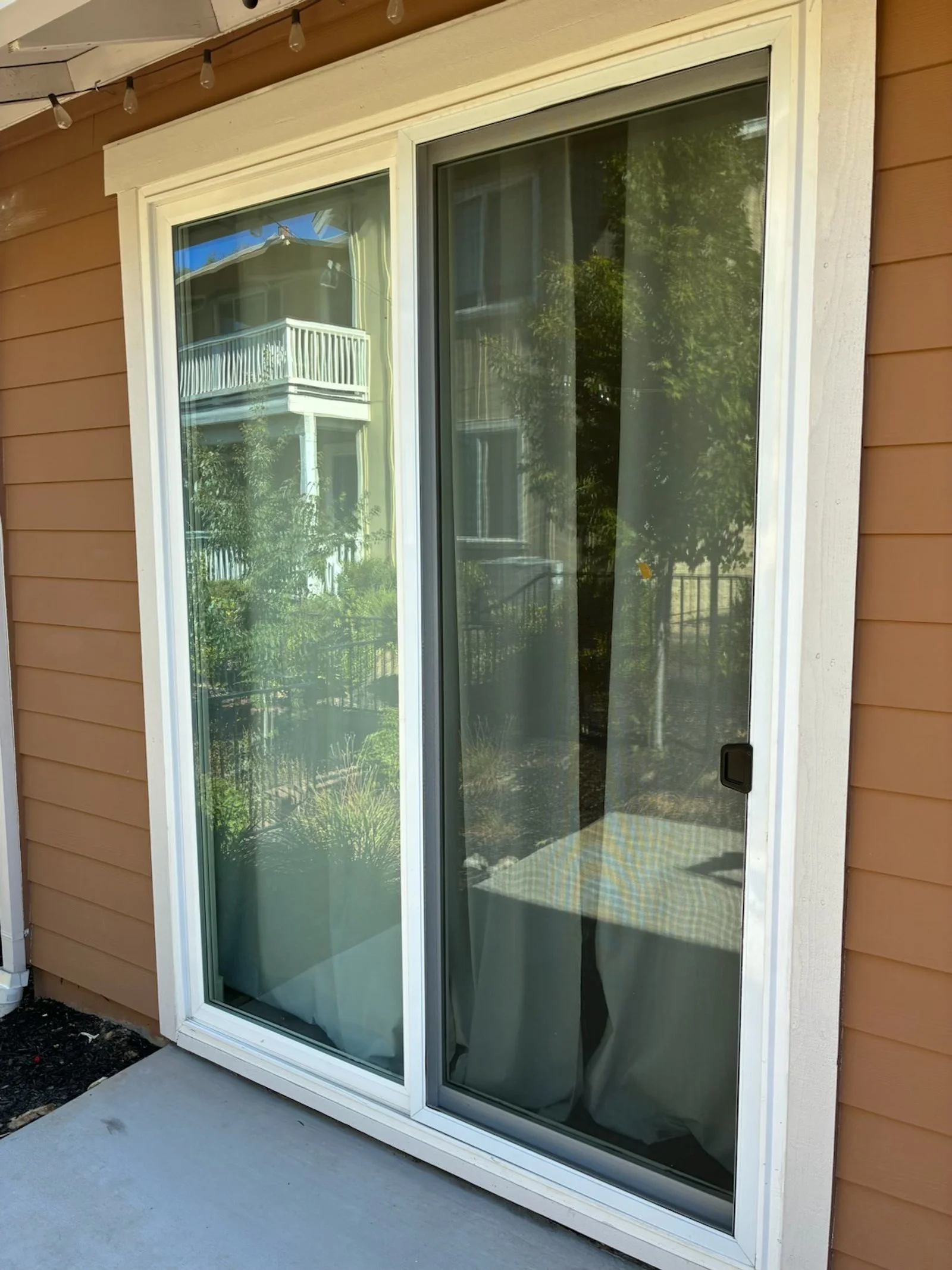 Sliding glass door with white frame on a brown exterior wall, reflecting a neighboring house with a balcony and green trees.