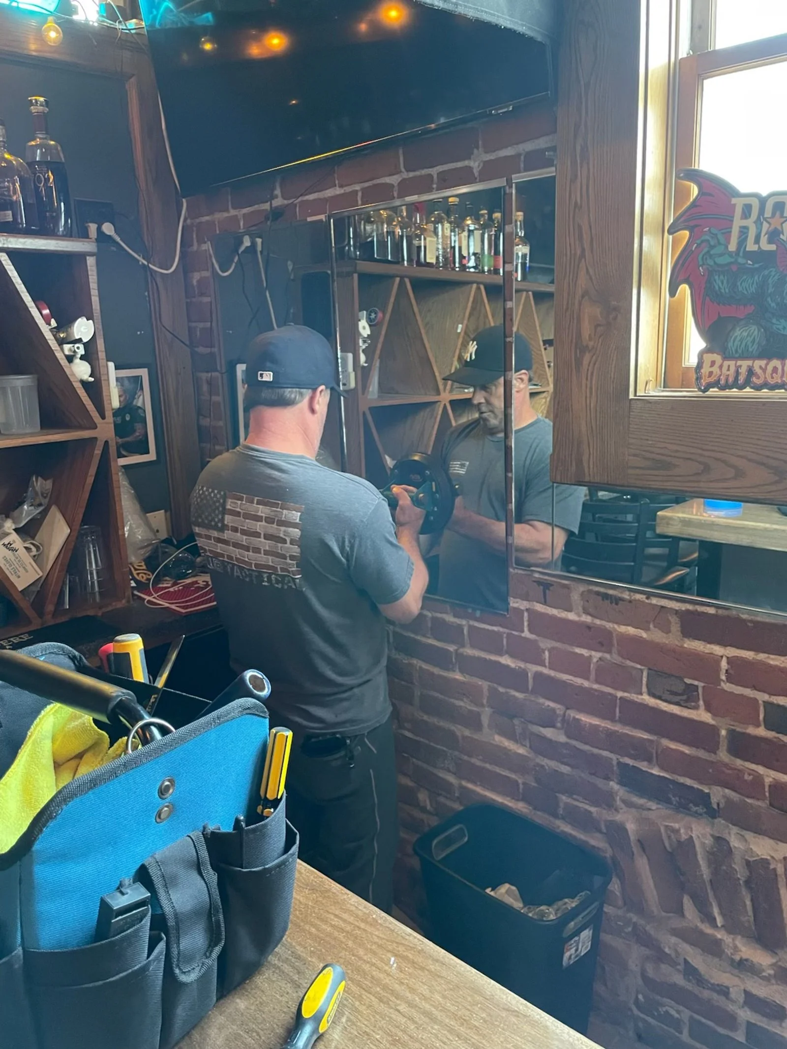 A man wearing a baseball cap and a gray t-shirt is looking at his reflection in a mirror, lifting a small dumbbell. The setting appears to be a bar or restaurant with brick walls, shelves with bottles, and a window with a sign that says "BATSQU" with