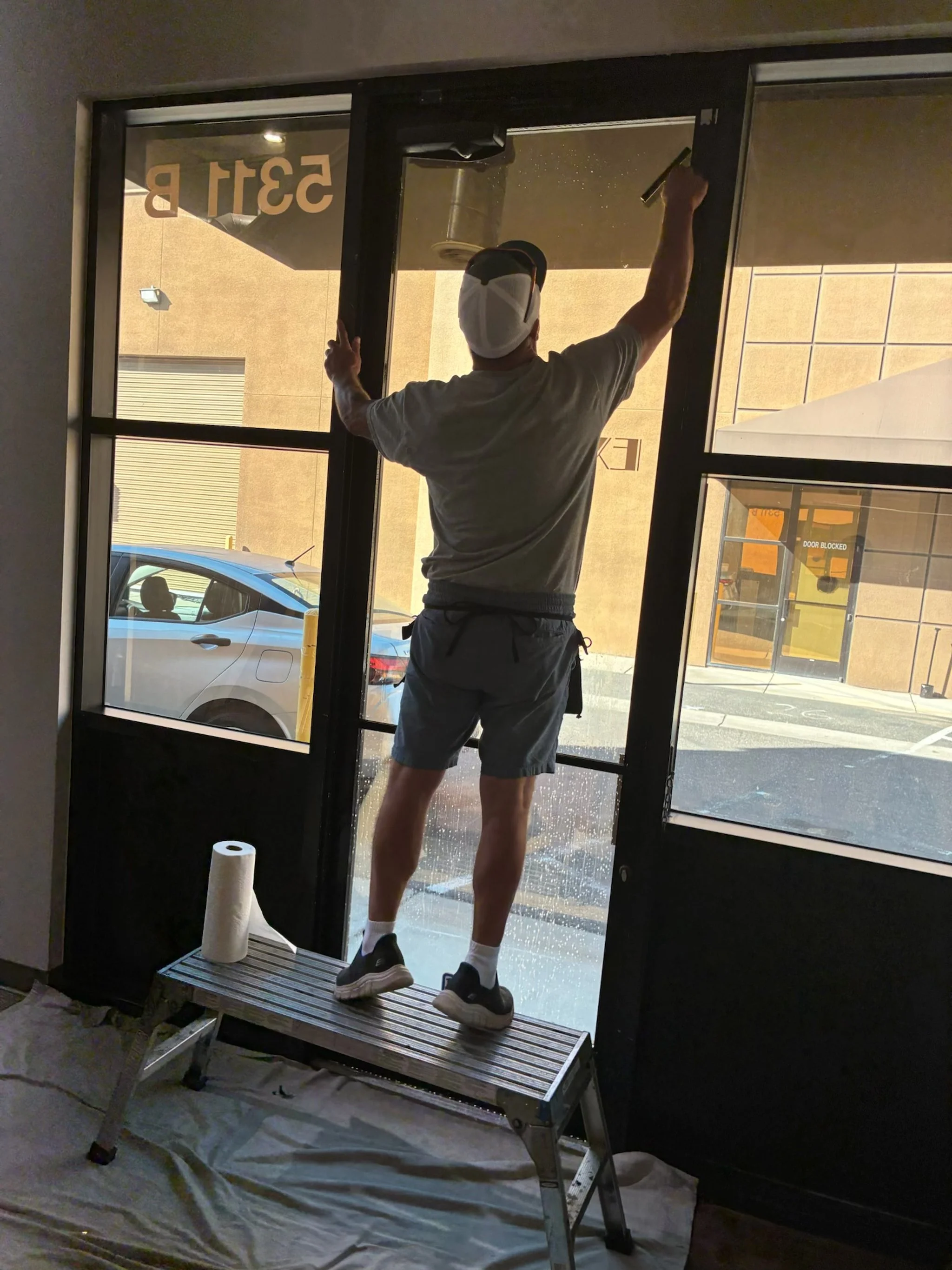 A man wearing a white cap, gray t-shirt, and shorts is cleaning a glass door from inside a building. He is standing on a metal step ladder and using a squeegee to wipe the glass. There is a roll of paper towels on a small tray attached to the ladder.