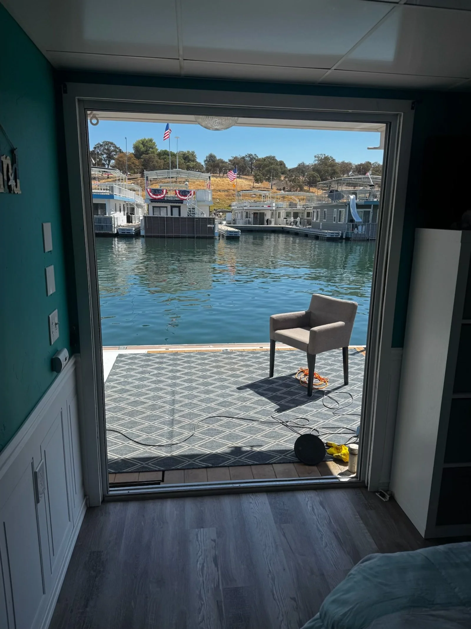 View from inside a house looking out through a sliding glass door at a marina with houseboats and American flags on a sunny day.
