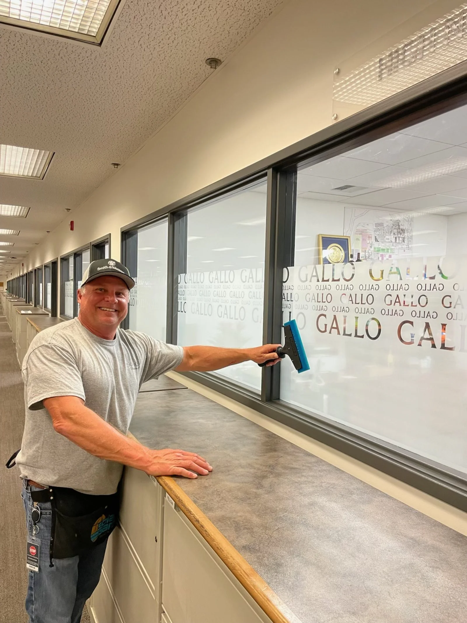 A man cleaning a glass window in an office hallway with a squeegee. The window has the words 'GALLO GALLO GALO' printed on it repeatedly.