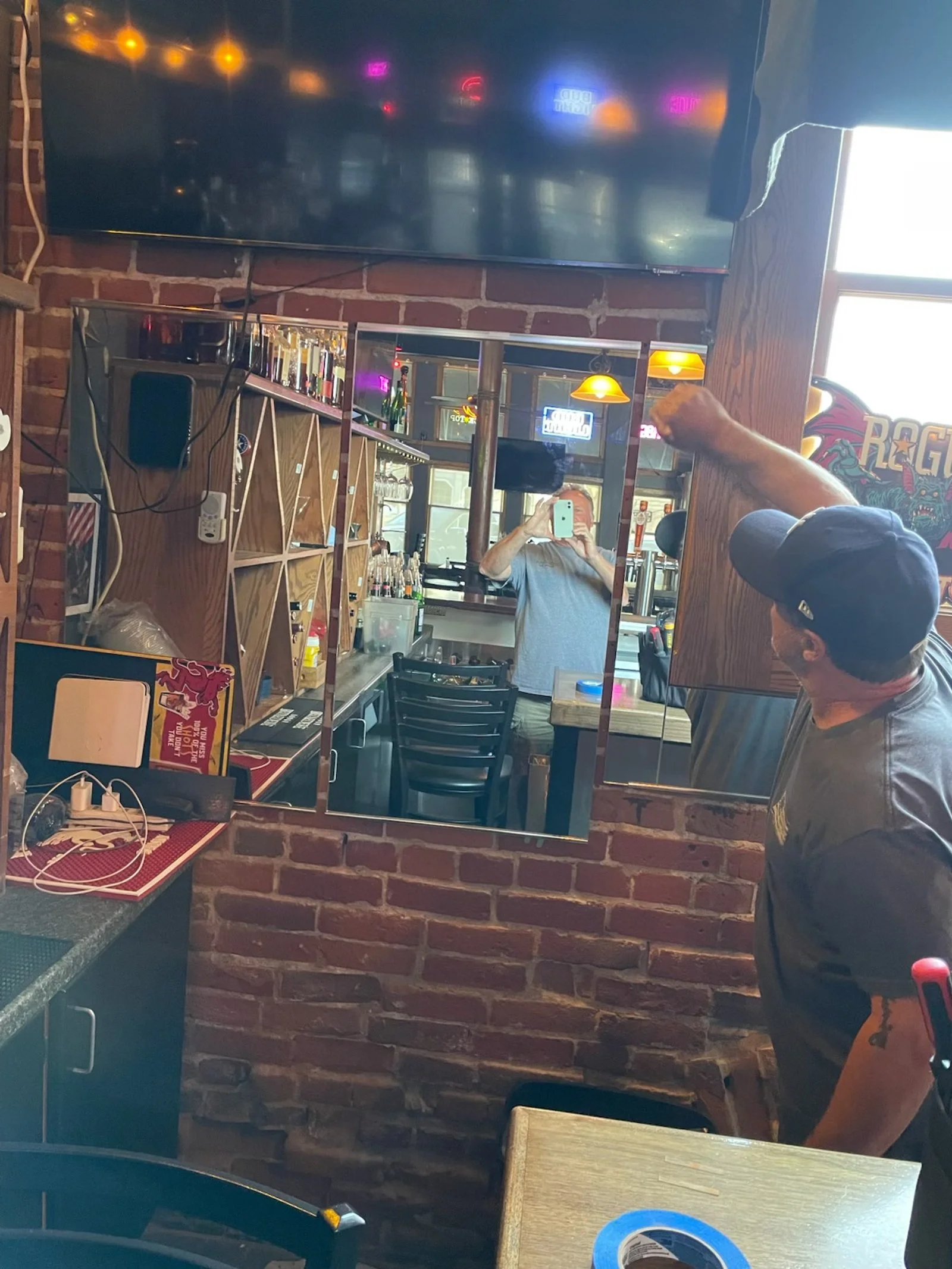 A man with a baseball cap and gray t-shirt raises his fist in a bar with exposed brick walls. There is a mirror behind him reflecting another man taking a selfie with a blue phone. The bar countertop has various bottles and glasses, and a large telev