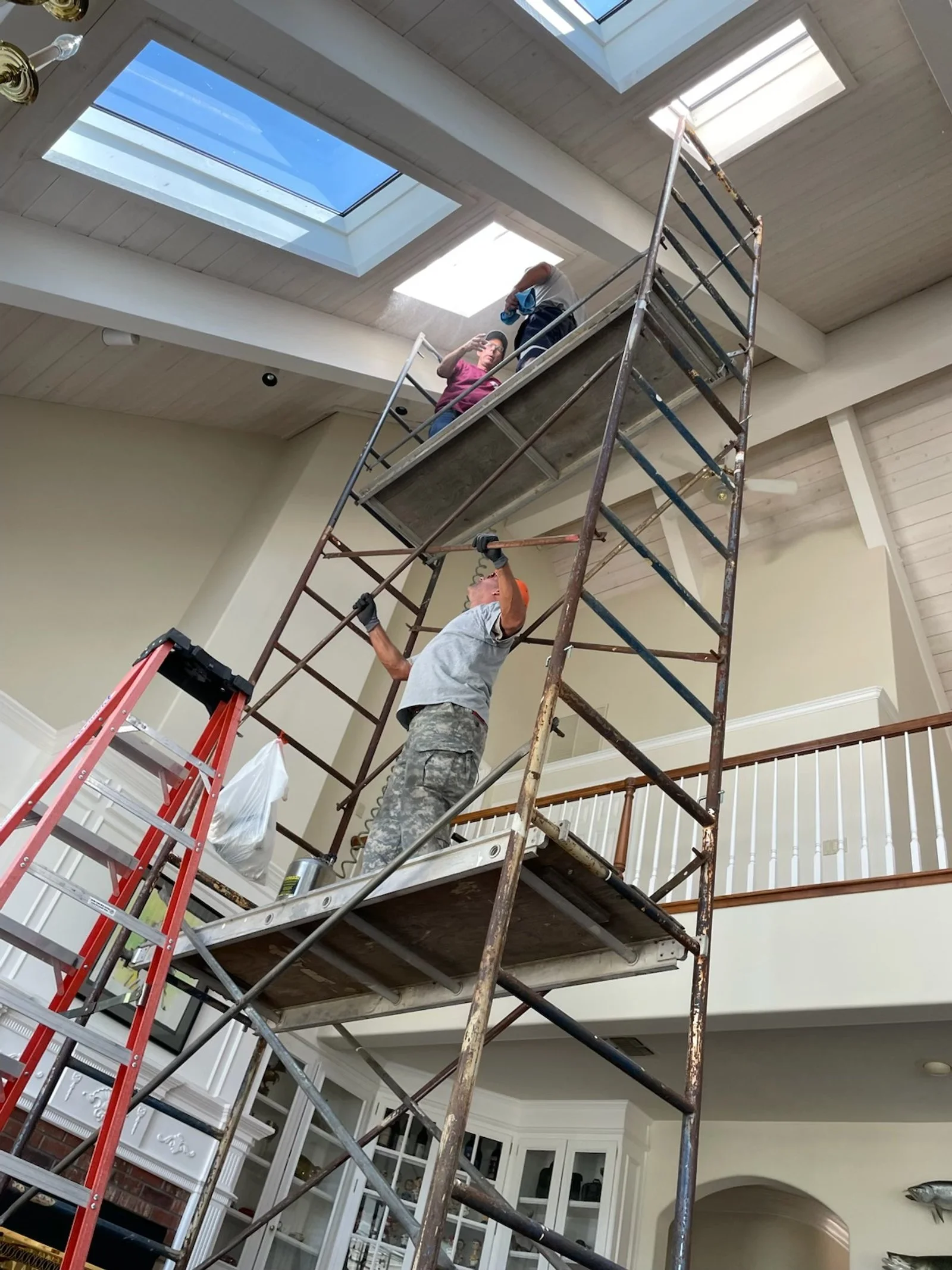 Two workers on a scaffold installing or repairing a skylight in a living room ceiling, with a ladder nearby.