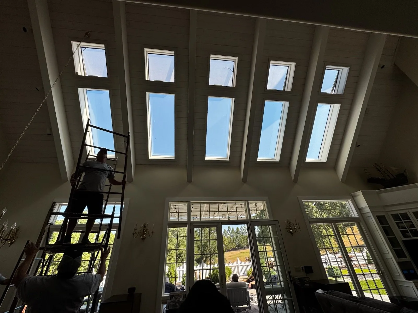 People installing or inspecting skylights in a high ceiling of a bright living room with large windows and outdoor view.
