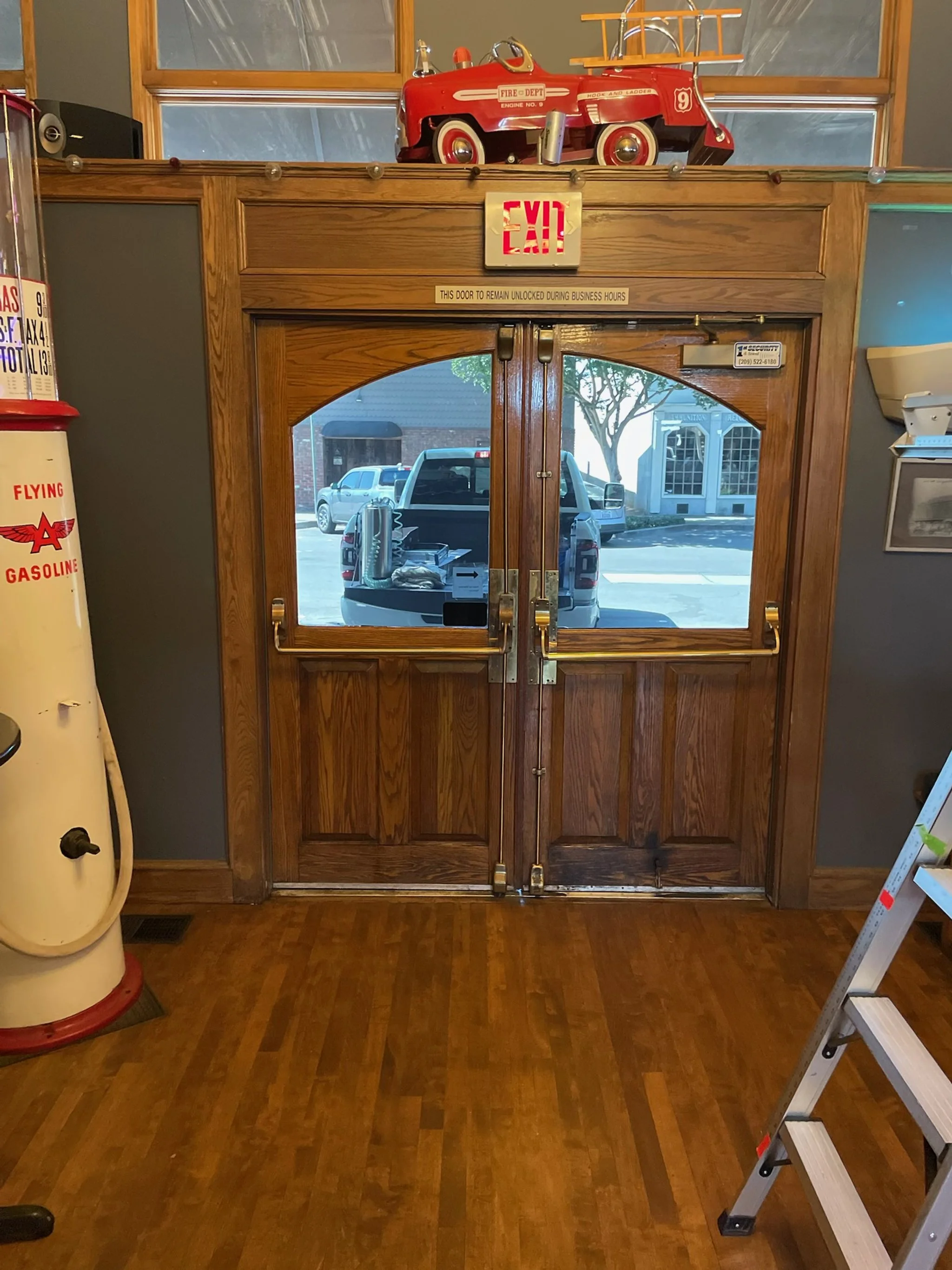 Wooden double doors with glass panels, located inside a building. Outside the doors, a parking lot with a truck and other cars is visible. A vintage fire truck model is placed on top of the doors, and an exit sign is mounted above them.