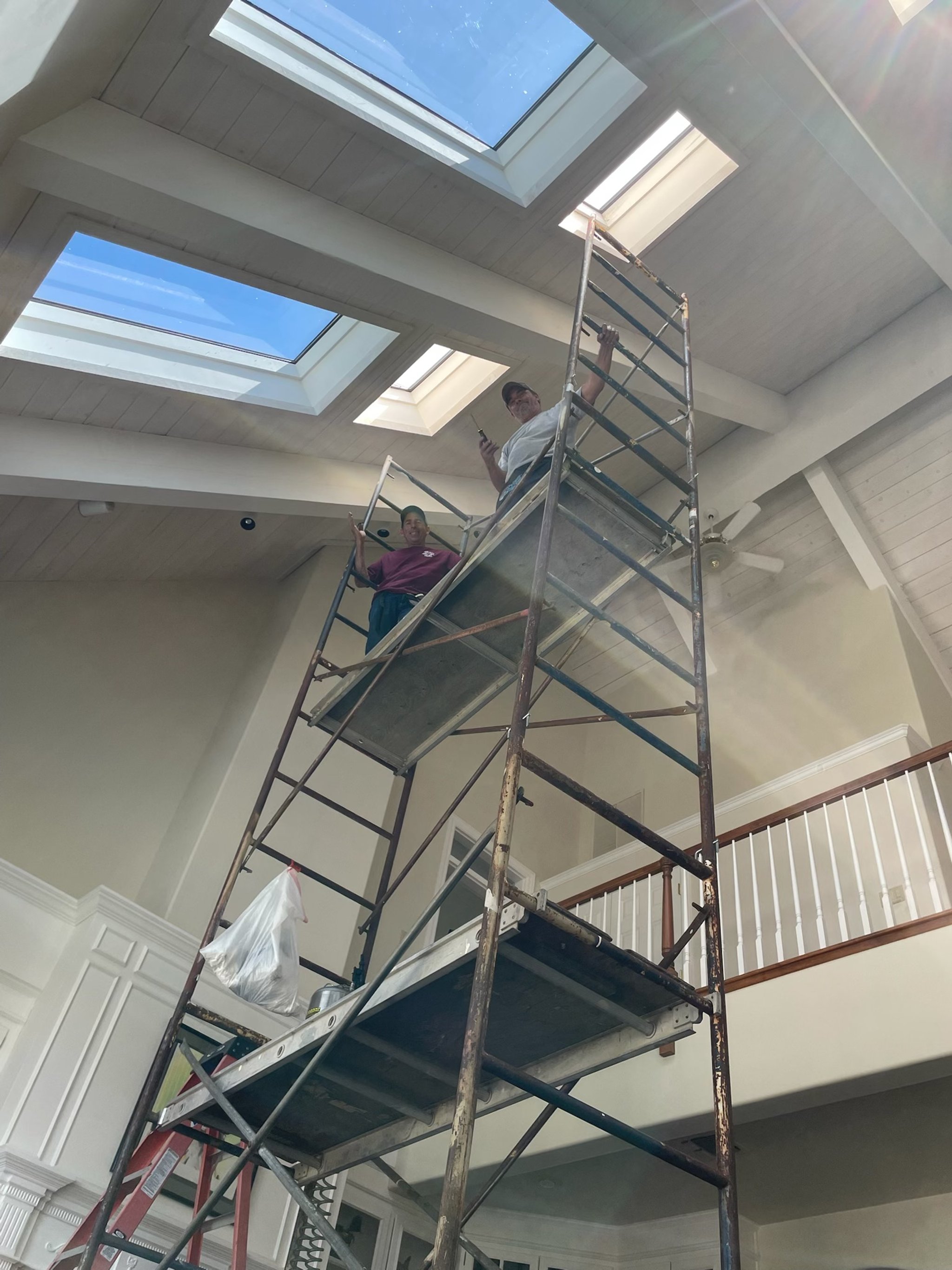 Two people on scaffolding inside a house with skylights in the ceiling, working on the roof. One person is holding a tool and looking down, while the other is smiling and looking at the camera.