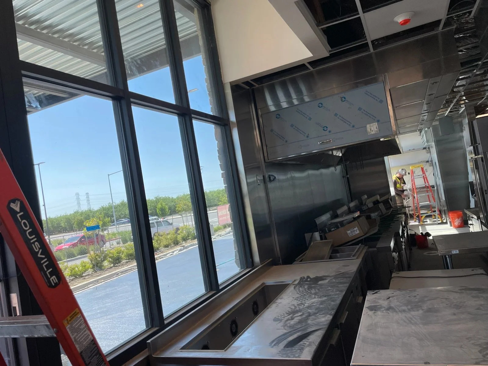 Commercial kitchen under construction with stainless steel counters, construction worker, ladder, and large window showing parking lot outside.