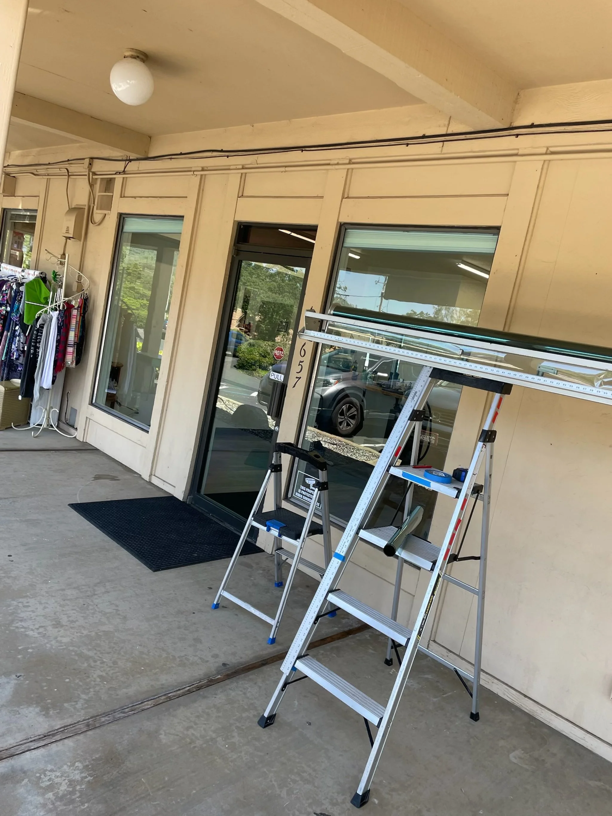 Outside of a storefront with three large windows, a black door, and a black mat. Two step ladders with a long metal object on top are set up outside. To the left, a clothing rack with various garments is visible. The storefront has beige-colored wall