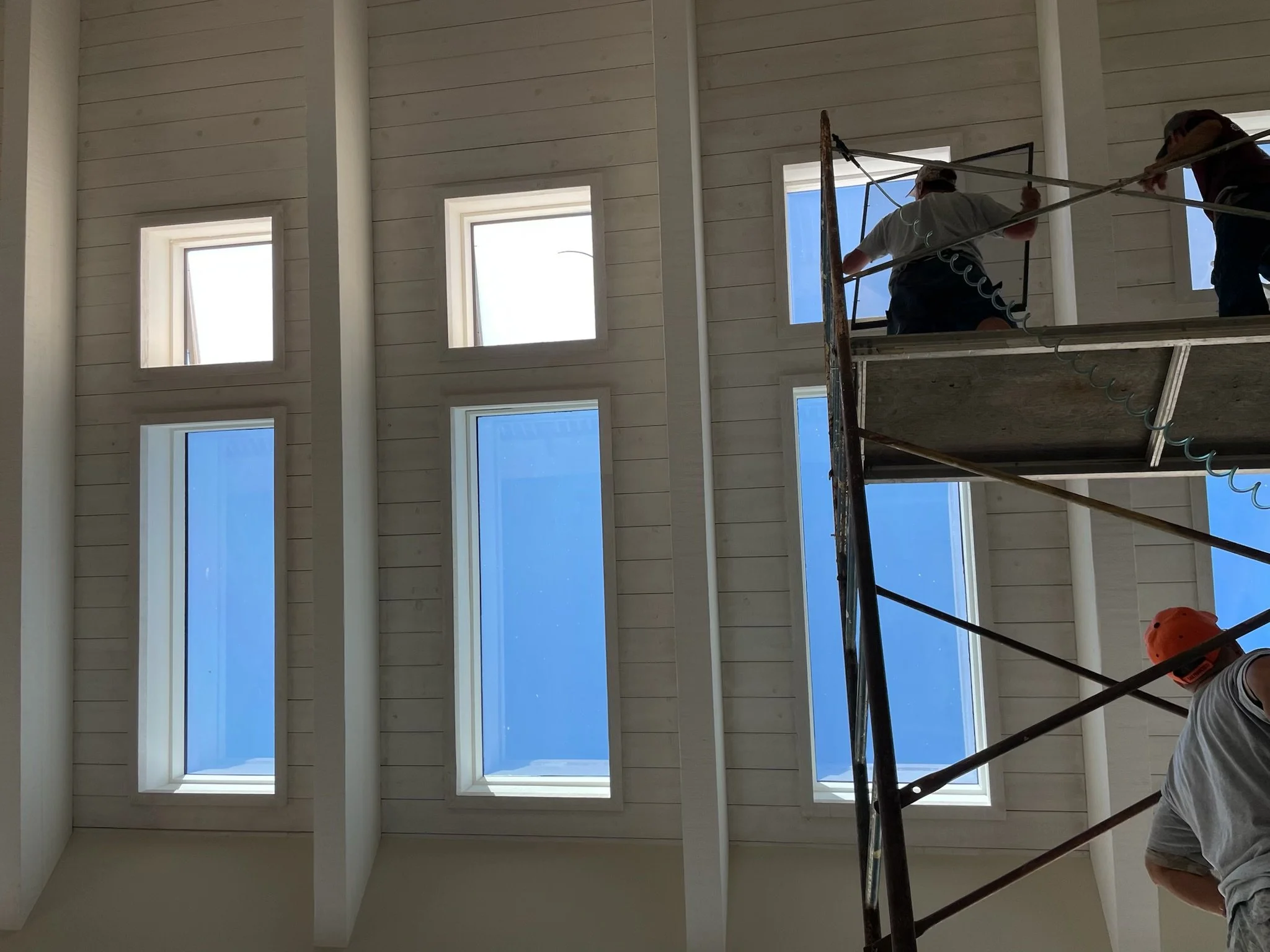 Construction workers on scaffolding installing windows in a building with a vaulted ceiling and multiple rectangular windows.