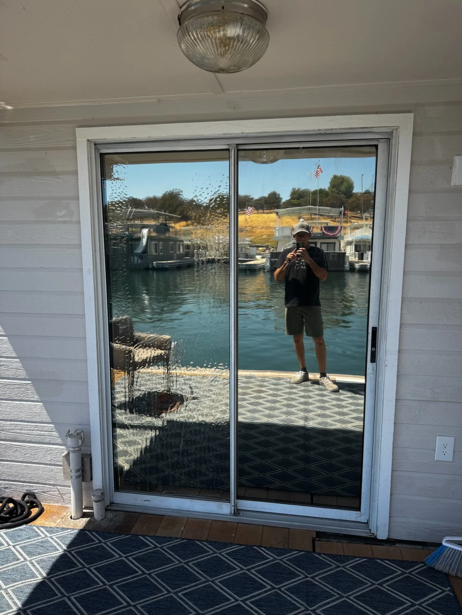 Reflection of a man taking a photo in a glass sliding door, with boats and flags in the water behind him.