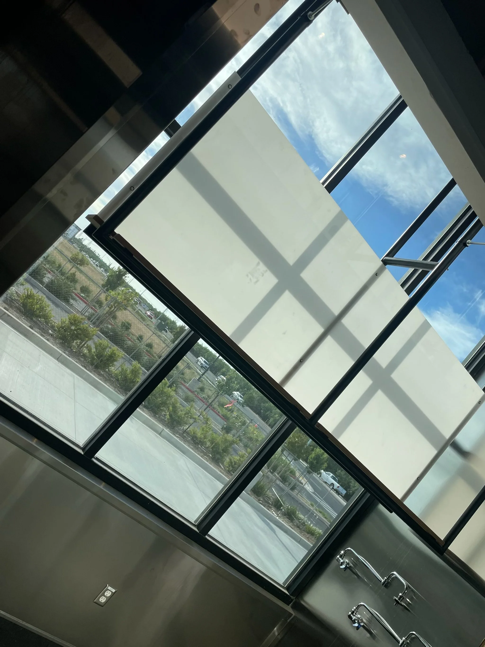 View of large windows with a view of a parking lot and cloudy blue sky outside, with part of an interior window frame and some metal fixtures visible.