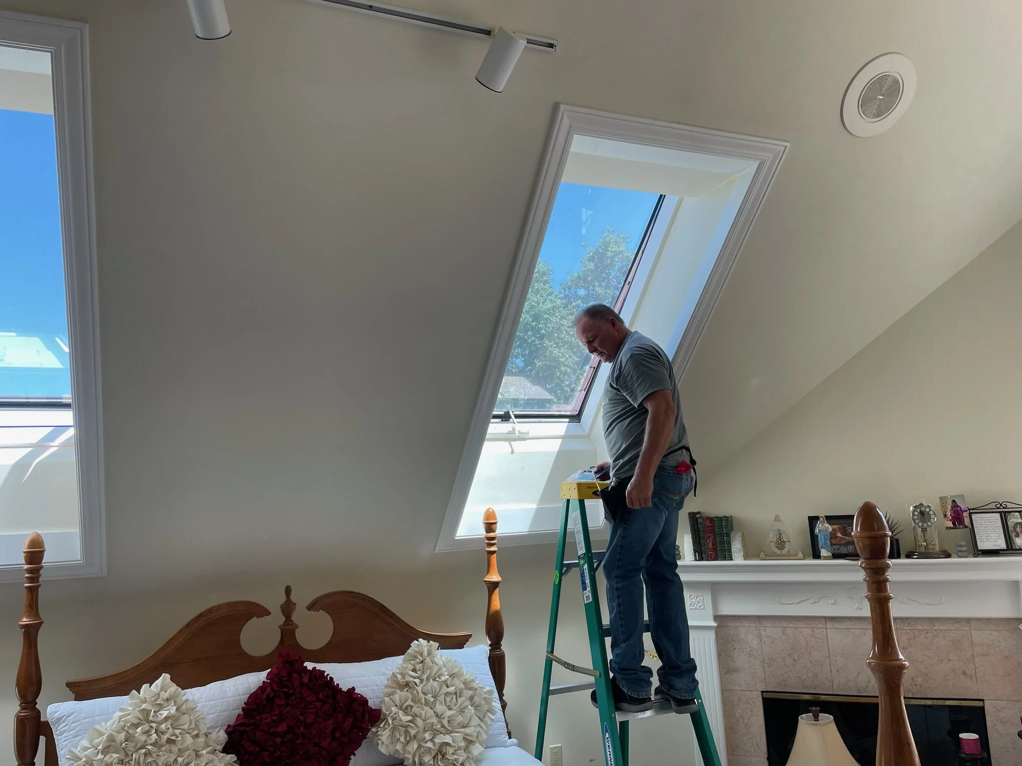 A man standing on a ladder installing or working on a skylight window in a room with sloped ceilings. The room has a bed with decorative pillows and a fireplace with various framed photographs and decorations on the mantel.