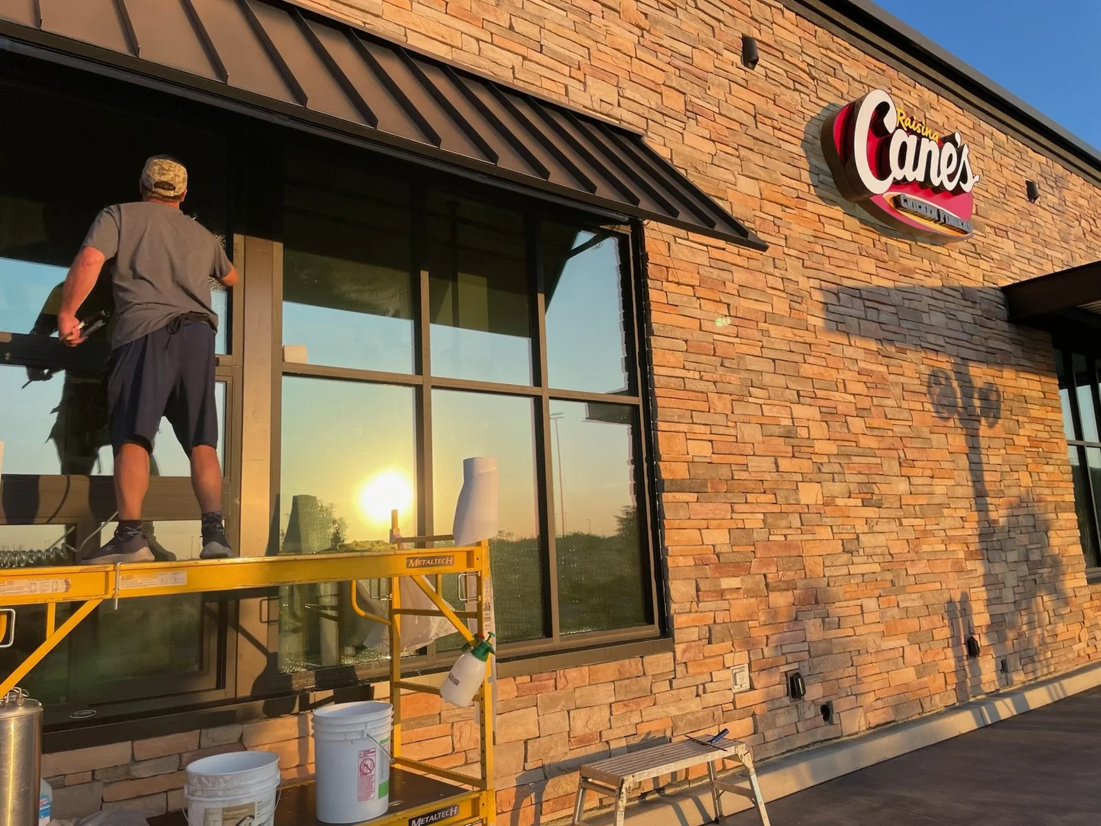 A worker on a yellow lift installing a large window at Raising Cane's restaurant during sunset, with the Cane's sign visible on the brick exterior wall.