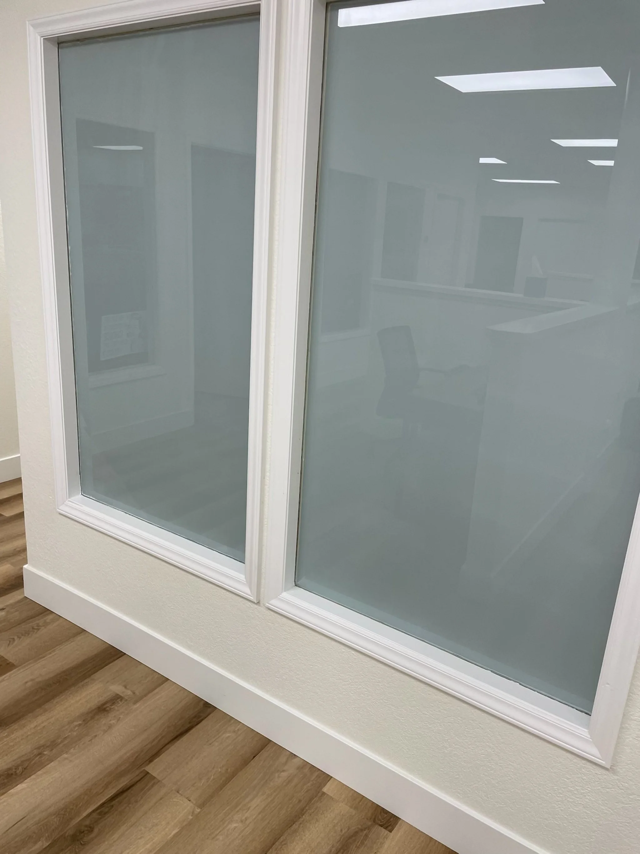 Interior view of a window with frosted glass and white frame, showing reflection of office chairs and lights inside an office.