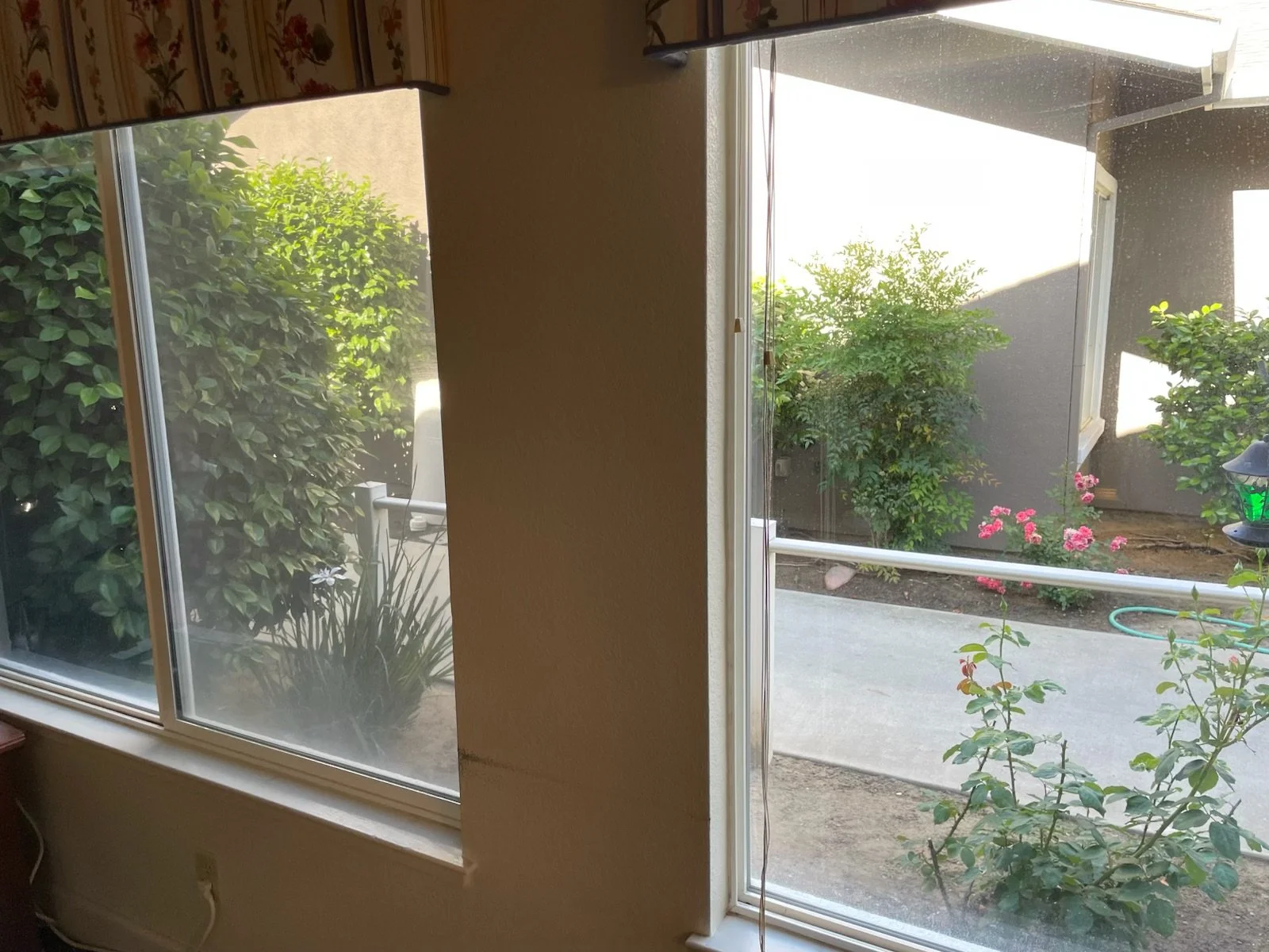View through two side-by-side house windows showing a small garden with bushes, pink flowers, a cement walkway, and a grey house with a window in the background.