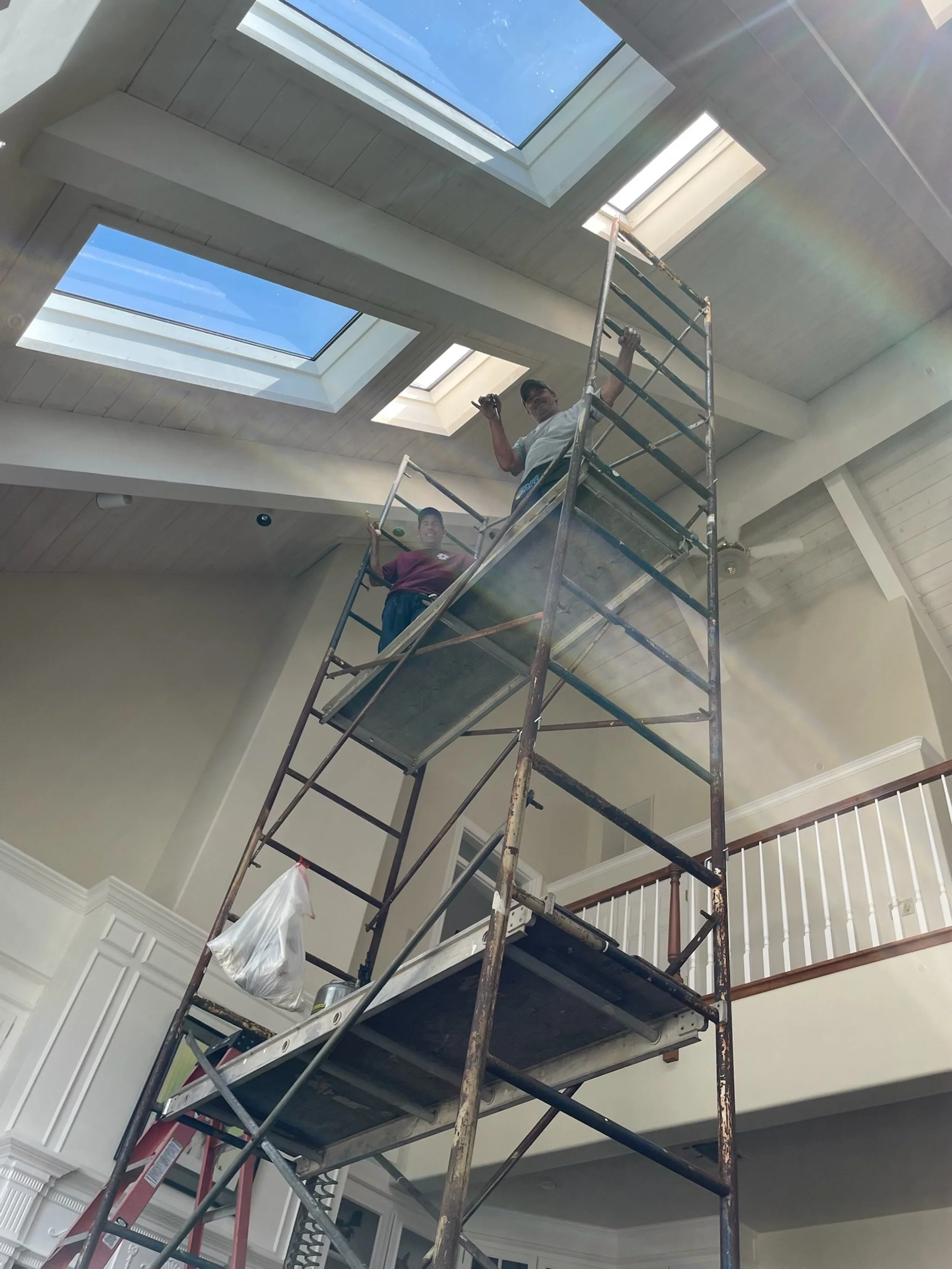 Two workers standing on scaffolding inside a house, working near a large skylight on the ceiling.