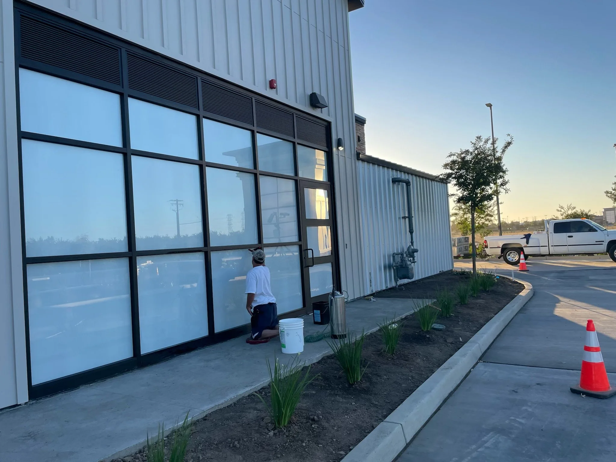 A person in a white shirt and dark shorts kneels in front of large glass windows outside a commercial building, which has a metal exterior and some small plants along the curb, with orange traffic cones nearby, during daylight.