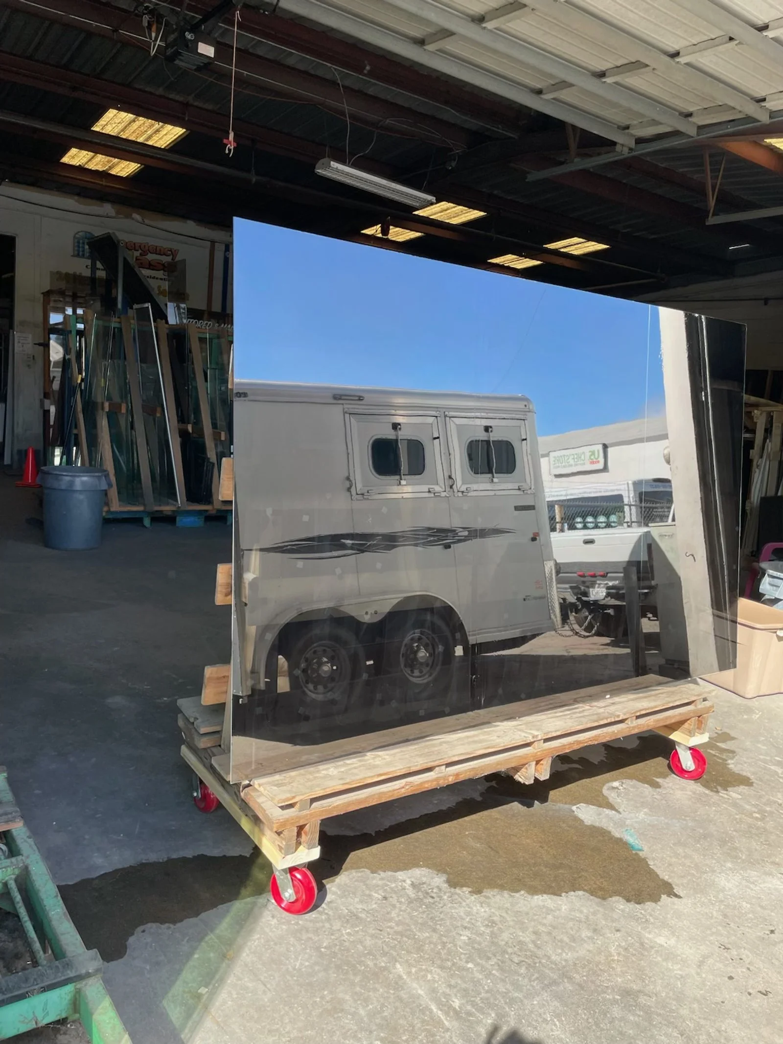 A large mirror reflecting the image of a white trailer with two small windows, a truck, and a blue sky, placed on a wooden platform with red wheels inside an industrial warehouse.