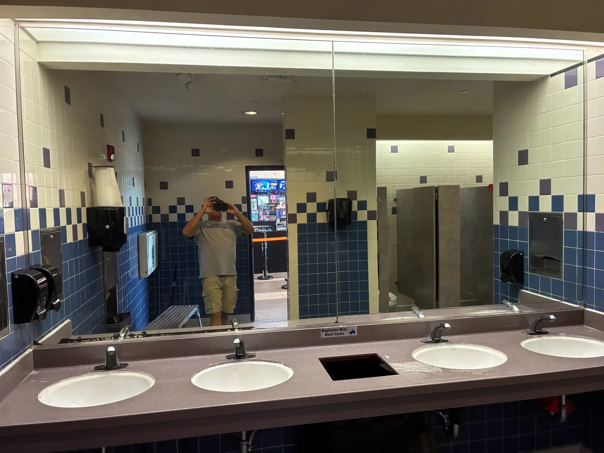 Public bathroom with four sinks, large mirror, soap dispensers, and a person taking a photo.