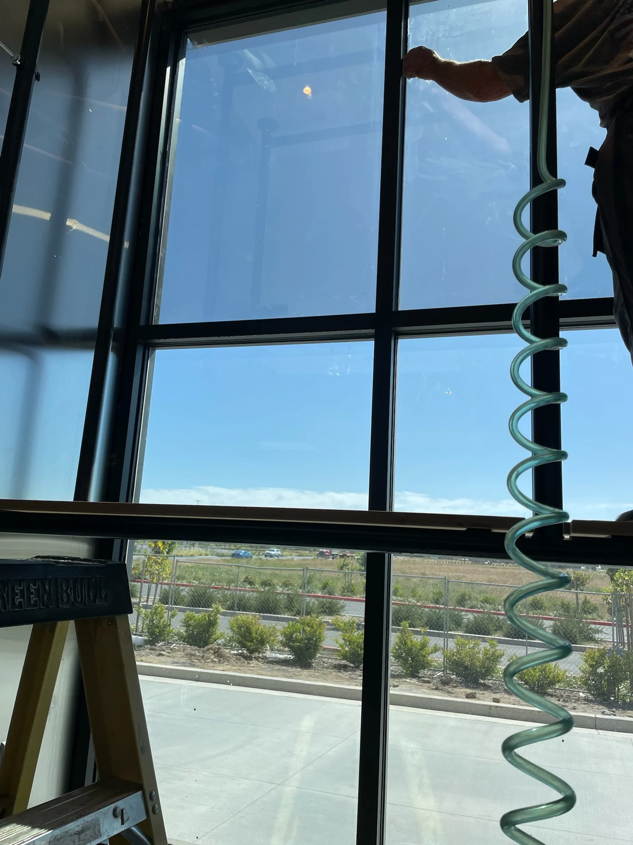 View through large glass windows showing a blue sky, some clouds, and parked cars outside. A person's arm and hand are visible, and a green spiral object is hanging down inside. Part of a wooden ladder with the words "GREEN BUILD" on it is in the low
