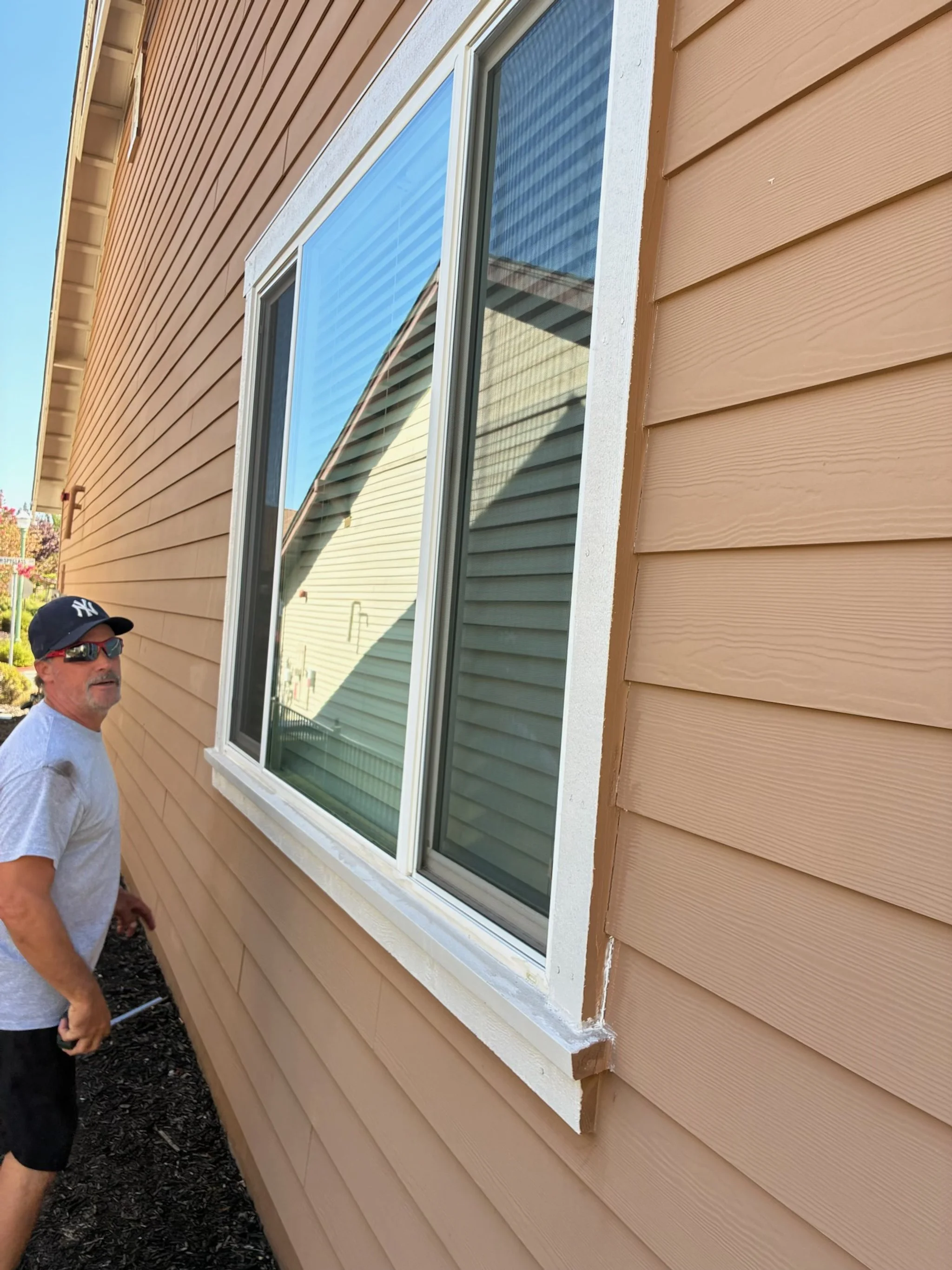 A man standing outside a house with tan horizontal siding, wearing sunglasses, a dark baseball cap, a light gray T-shirt, and black shorts, next to a large window with white trim.