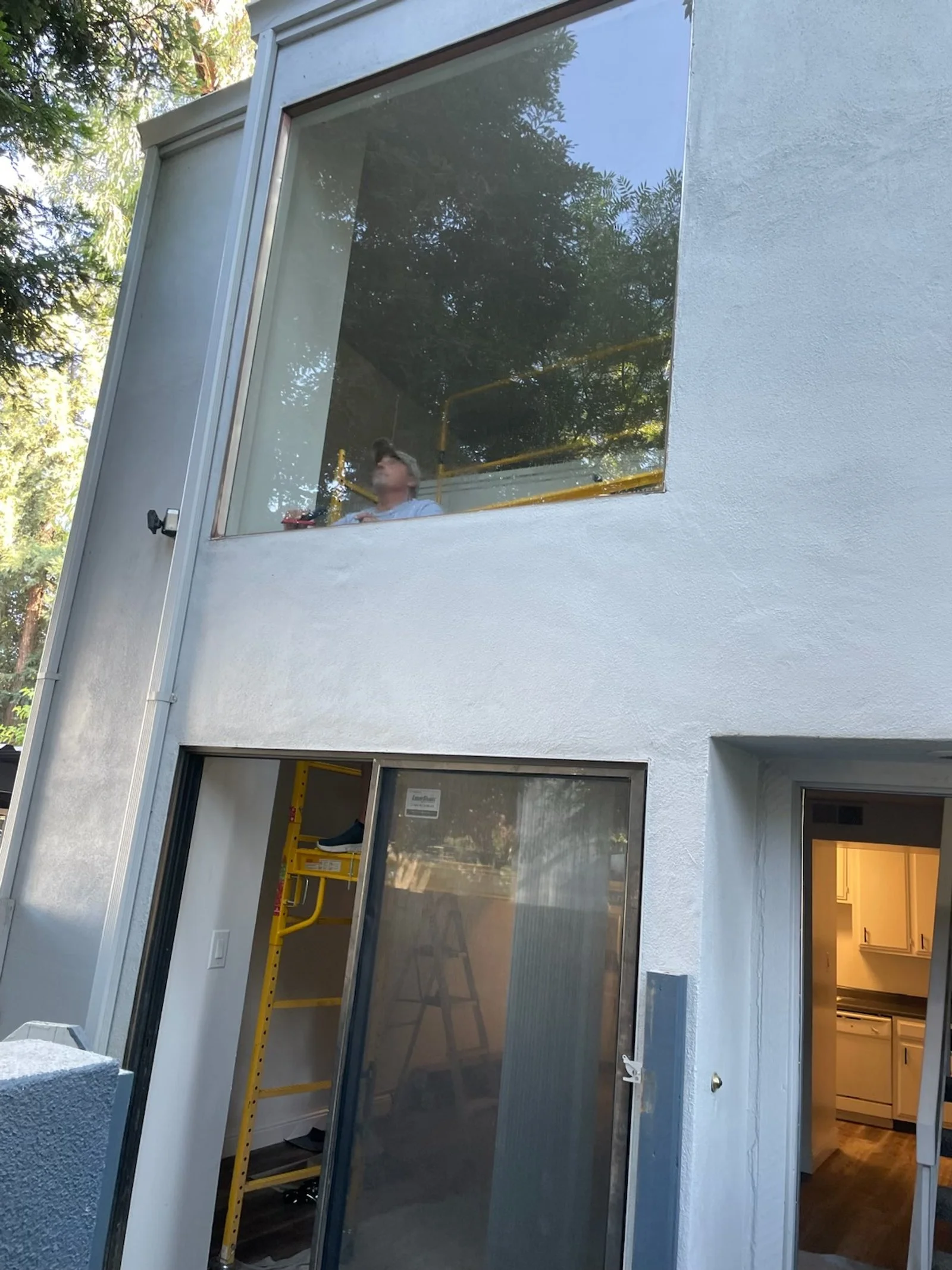 View of a two-story house with large windows. The upper window shows a person working or sitting inside, while the lower part shows a sliding glass door with a yellow ladder visible through it, leading to the second floor. The interior includes a vis