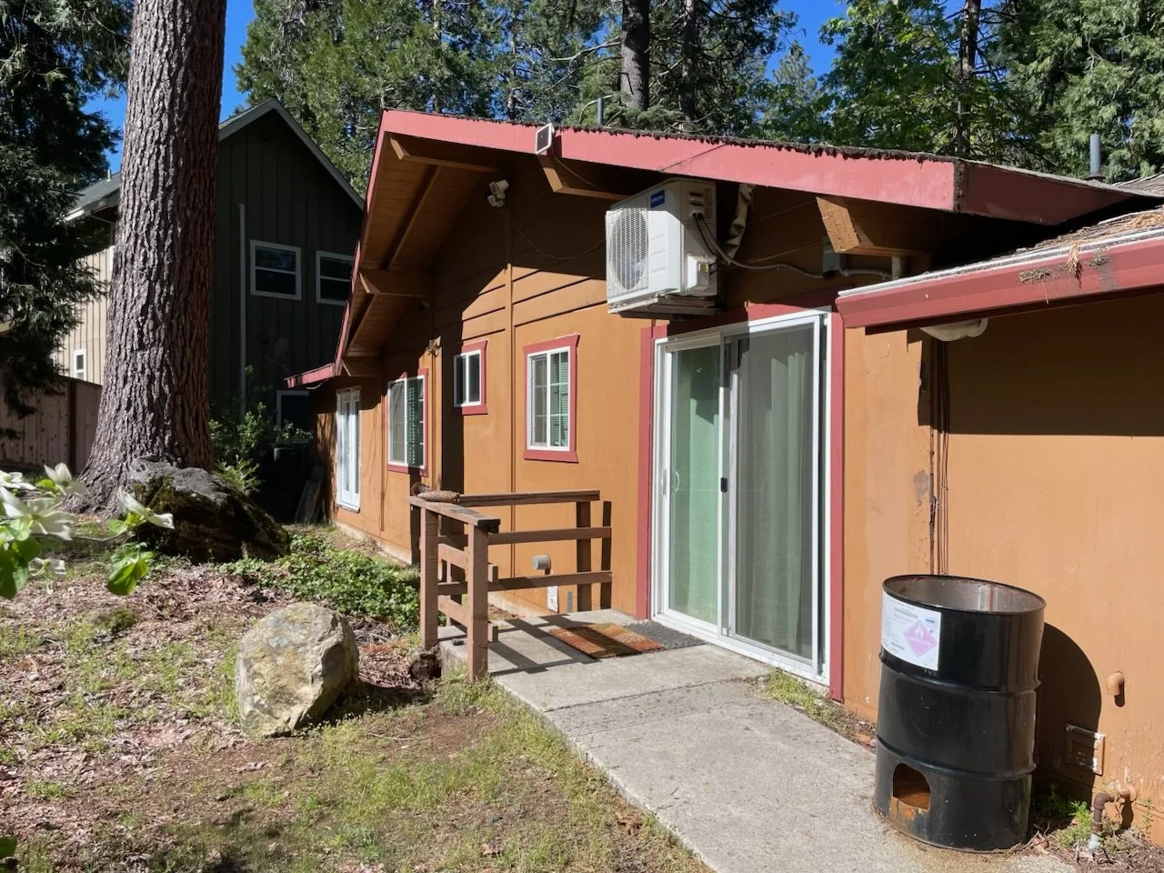 Backyard view of a brown house with a red roof and white sliding glass door, a small wooden ramp, an air conditioning unit, and a black barrel-shaped container on a concrete pad, surrounded by trees and plants.