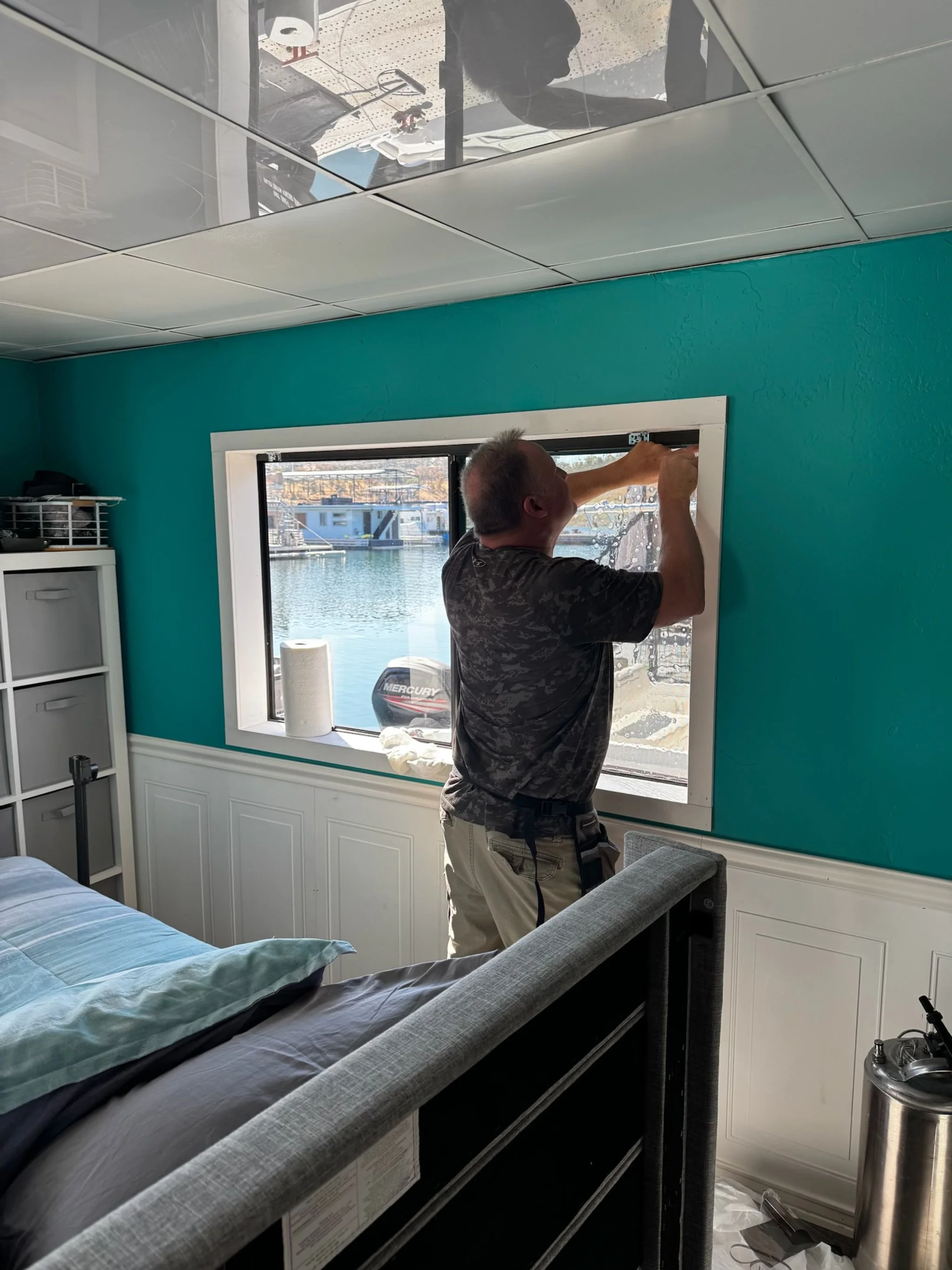 A man installing or fixing a white window frame in a turquoise wall in a bedroom with a bed, a metal storage unit, and a view of water and boats outside.
