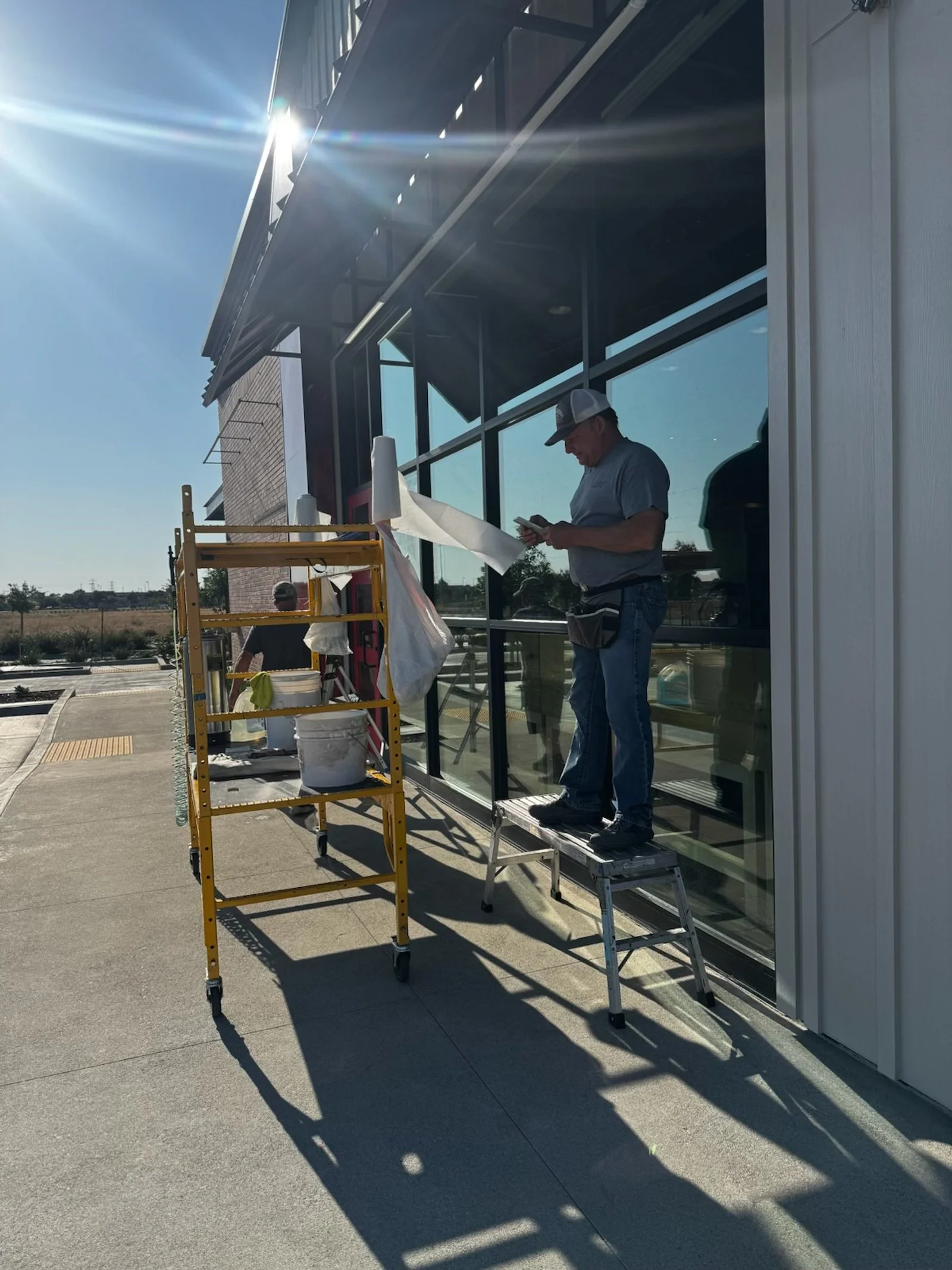 A man standing on a small ladder painting or printing a large window outside a modern commercial building, with a scaffolding nearby, on a sunny day.