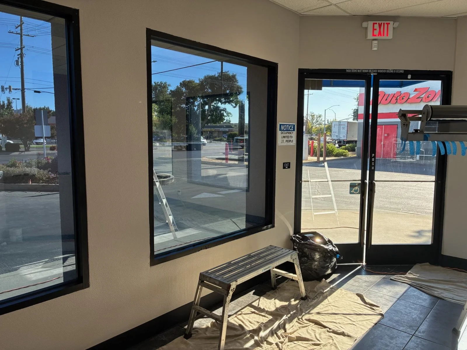 Interior of a store with large front windows and glass door, a metal step ladder, a black trash bag, and cloths on the floor. Outside there is a parking lot, trees, and a building with a red and white sign that says 'Auto Zone'.