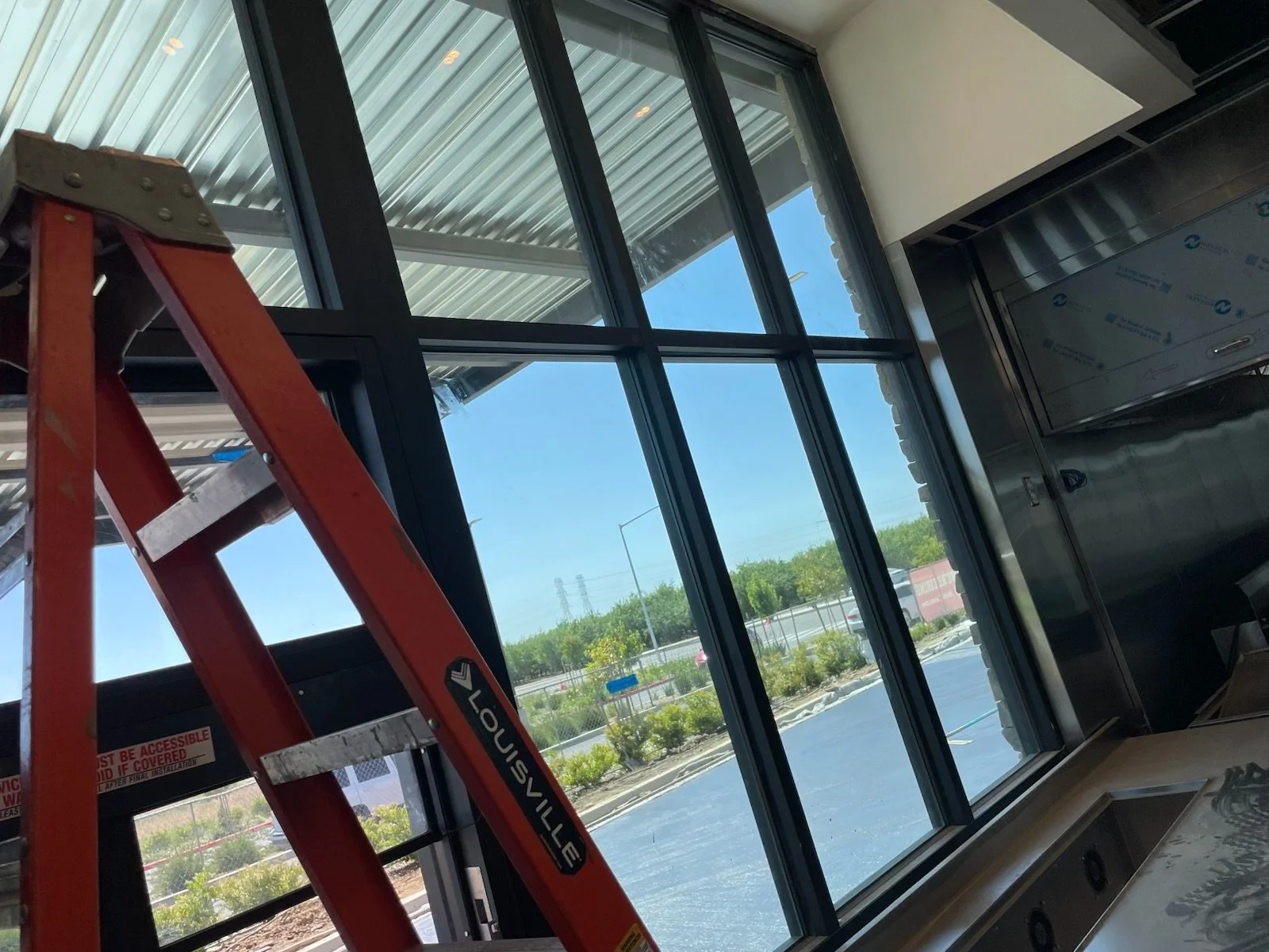 Interior of a building under construction, showing large glass windows, a red ladder, and metallic wall panels.