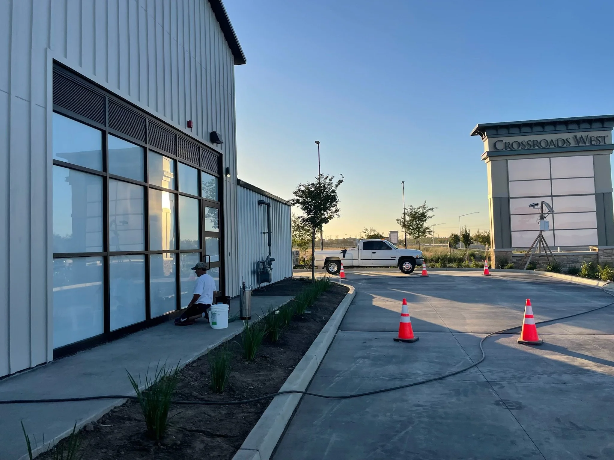 Construction worker kneeling outside a commercial building with large glass windows, working on the sidewalk during sunset. A white pickup truck is parked in the background near traffic cones. The area has some small plants lining the sidewalk, and a