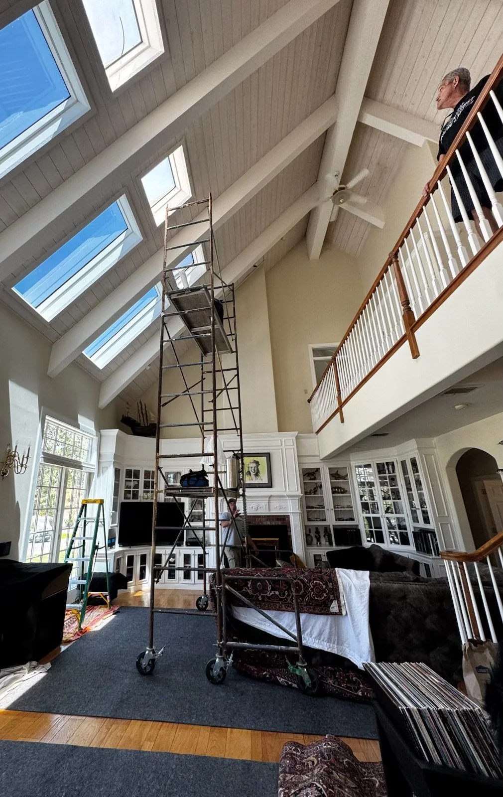 Ladder and scaffolding in a living room with high vaulted ceiling and skylights, person standing on the upper level, and a person taking a photo in front of a fireplace surrounded by built-in bookshelves.